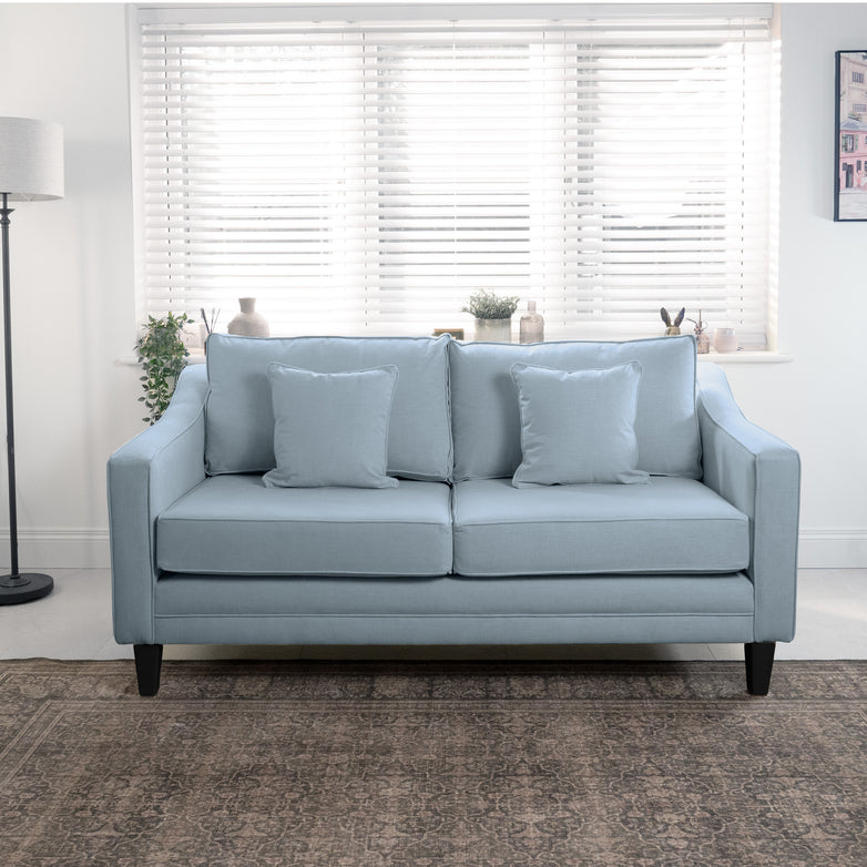 The Wiltshire 2 Seat Sofa in Sky Blue, handmade in England, sits gracefully in a bright room with cushions, alongside a sleek floor lamp and elegant blinds, adding charm and sophistication to any living space.