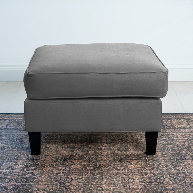 The Wiltshire Footstool in Smoke Grey, featuring dark legs, rests elegantly on a patterned rug by a white wall, showcasing modern appeal.