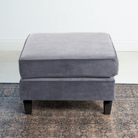 The versatile Wiltshire Velvet Footstool in Steel Grey sits elegantly on a patterned rug against a light wall backdrop.