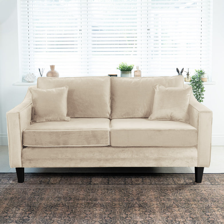 The Wiltshire 2 Seat Velvet Sofa in Cream, featuring two cushions, is placed before a window with white blinds and potted plants.