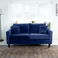 The Wiltshire 2 Seat Velvet Sofa in Royal Blue, complete with cushions, is elegantly placed by a window with blinds, surrounded by lush potted plants and a sleek floor lamp.