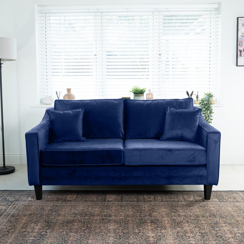 The Wiltshire 2 Seat Velvet Sofa in Royal Blue, complete with cushions, is elegantly placed by a window with blinds, surrounded by lush potted plants and a sleek floor lamp.