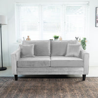 The Wiltshire 2 Seat Velvet Sofa in Feather Grey, featuring two cushions, is beautifully set before a window with white blinds amidst chic decor and lush plants. Handcrafted in England for an elegant touch.