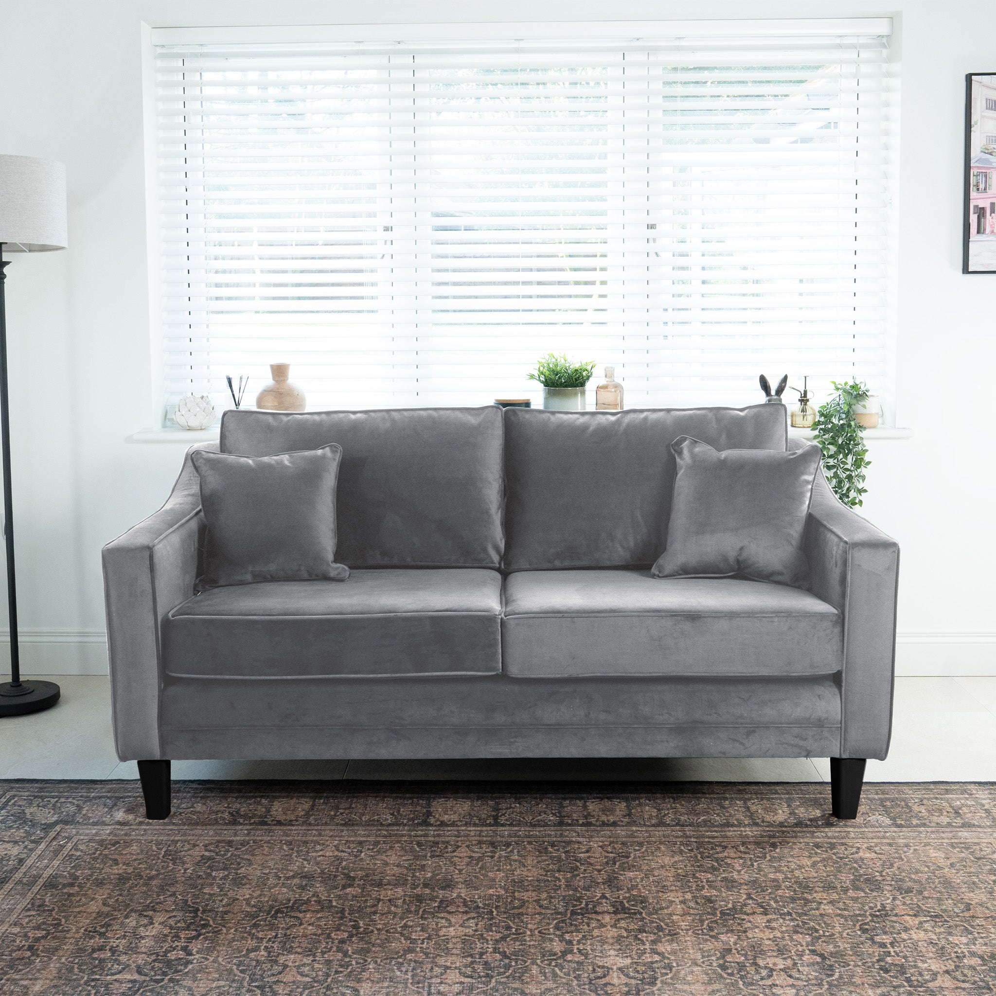 Wiltshire 2 Seat Velvet Sofa in Steel Grey with two cushions in a bright room, featuring a large window with blinds and potted plants on a shelf behind.
