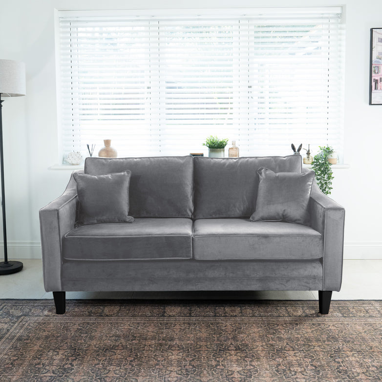 Wiltshire 2 Seat Velvet Sofa in Steel Grey with two cushions in a bright room, featuring a large window with blinds and potted plants on a shelf behind.