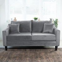 The Wiltshire 2 Seat Velvet Sofa in Steel Grey, handcrafted in England with two matching cushions, is placed before a window with blinds and potted plants.