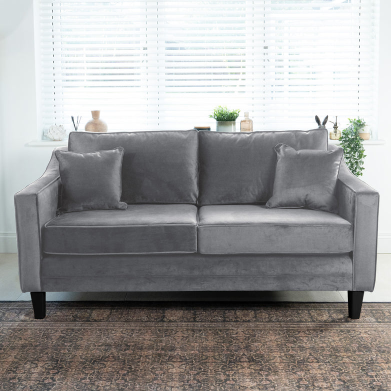 The Wiltshire 2 Seat Velvet Sofa in Steel Grey, handcrafted in England with two matching cushions, is placed before a window with blinds and potted plants.