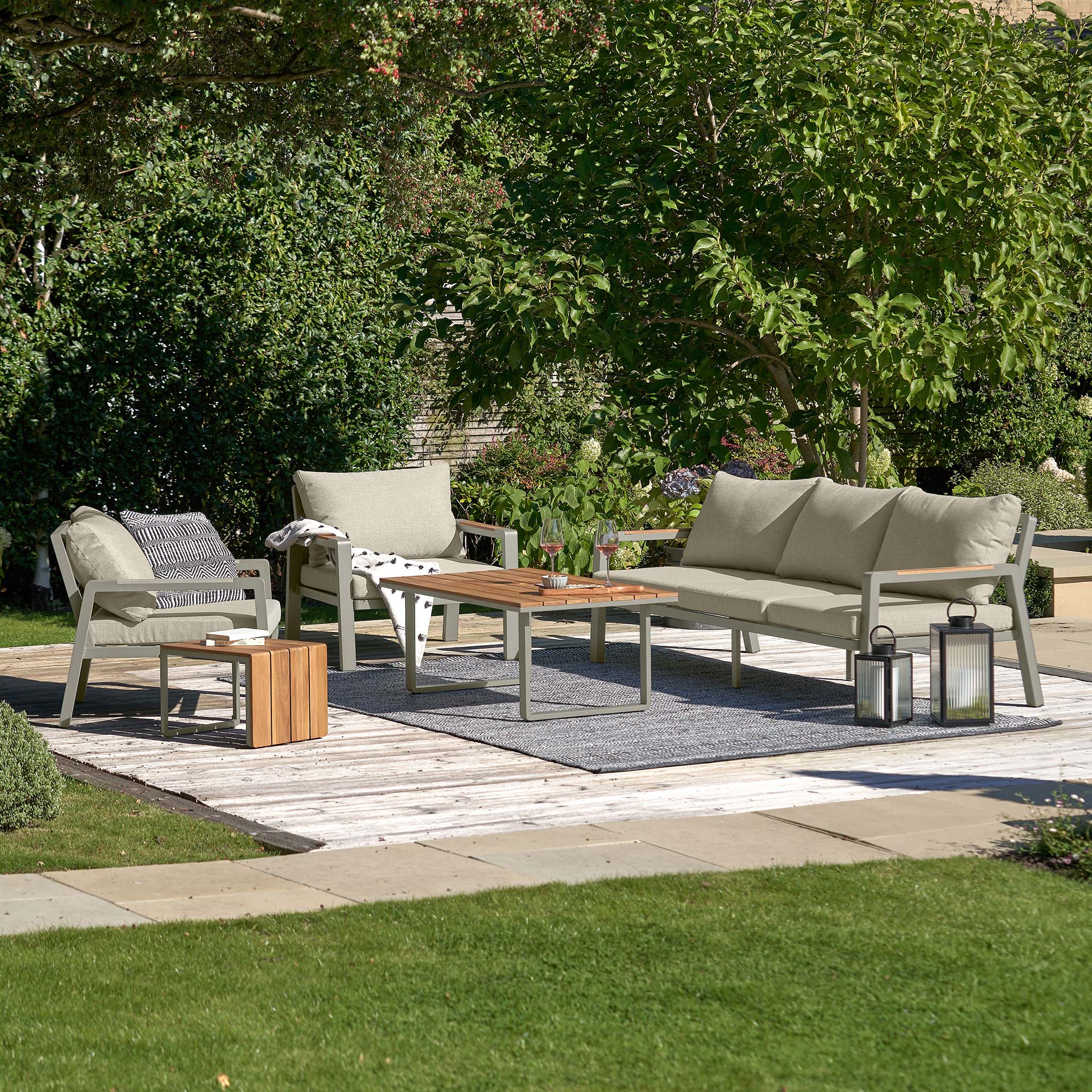 Lounge set with Pacific Lifestyle's Stockholm 3 Seat Sofa Set in Limestone, featuring a weatherproof sofa, chairs, and coffee table on a patio adorned with lanterns and a rug amidst lush greenery.