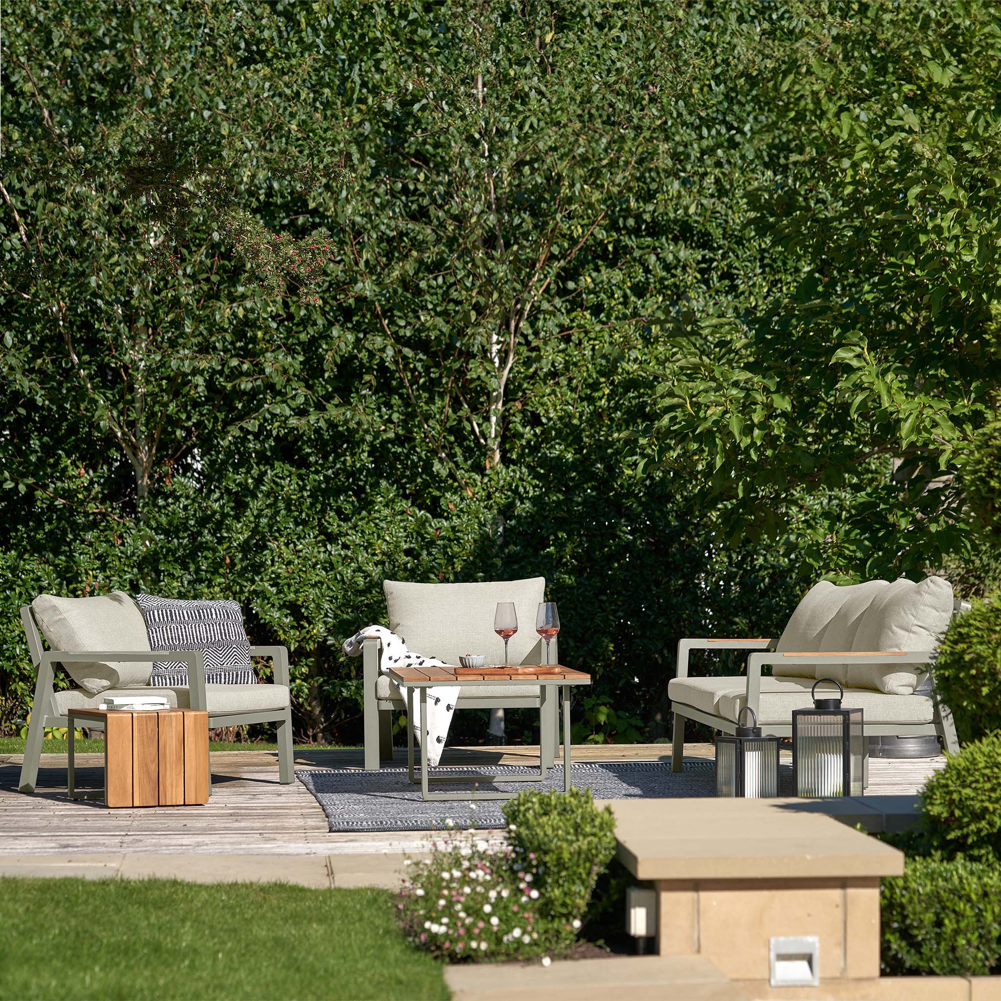 An outdoor patio showcases the Pacific Lifestyle Stockholm 3 Seat Sofa Set in Limestone with green cushioned seating and a teak wood table set with wine glasses, all amidst lush greenery.