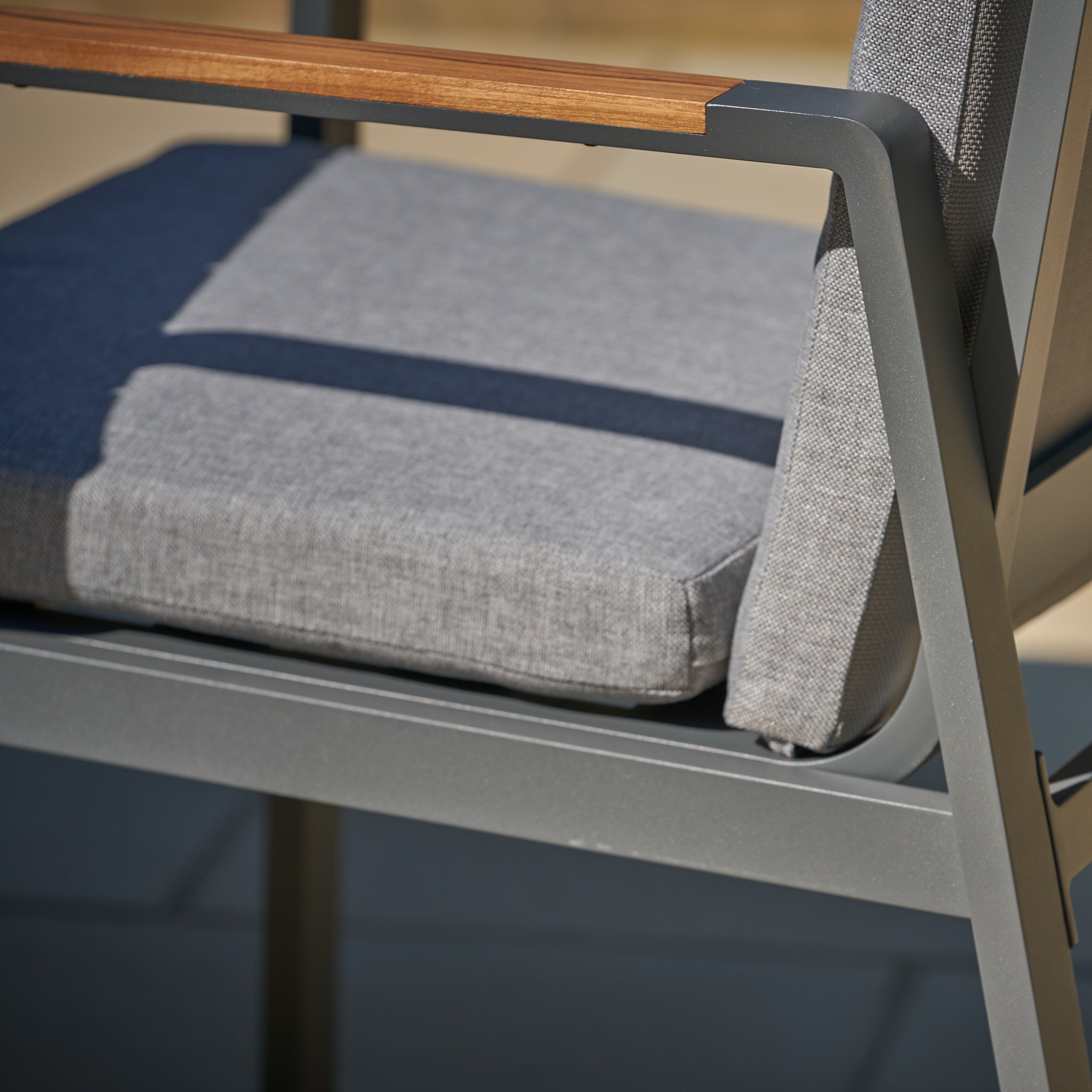Close-up of a Pacific Lifestyle Stockholm chair from the Marl Grey 6 Seat Dining Set, showcasing gray cushions, a metal frame, and teak wood armrests under sunlight.