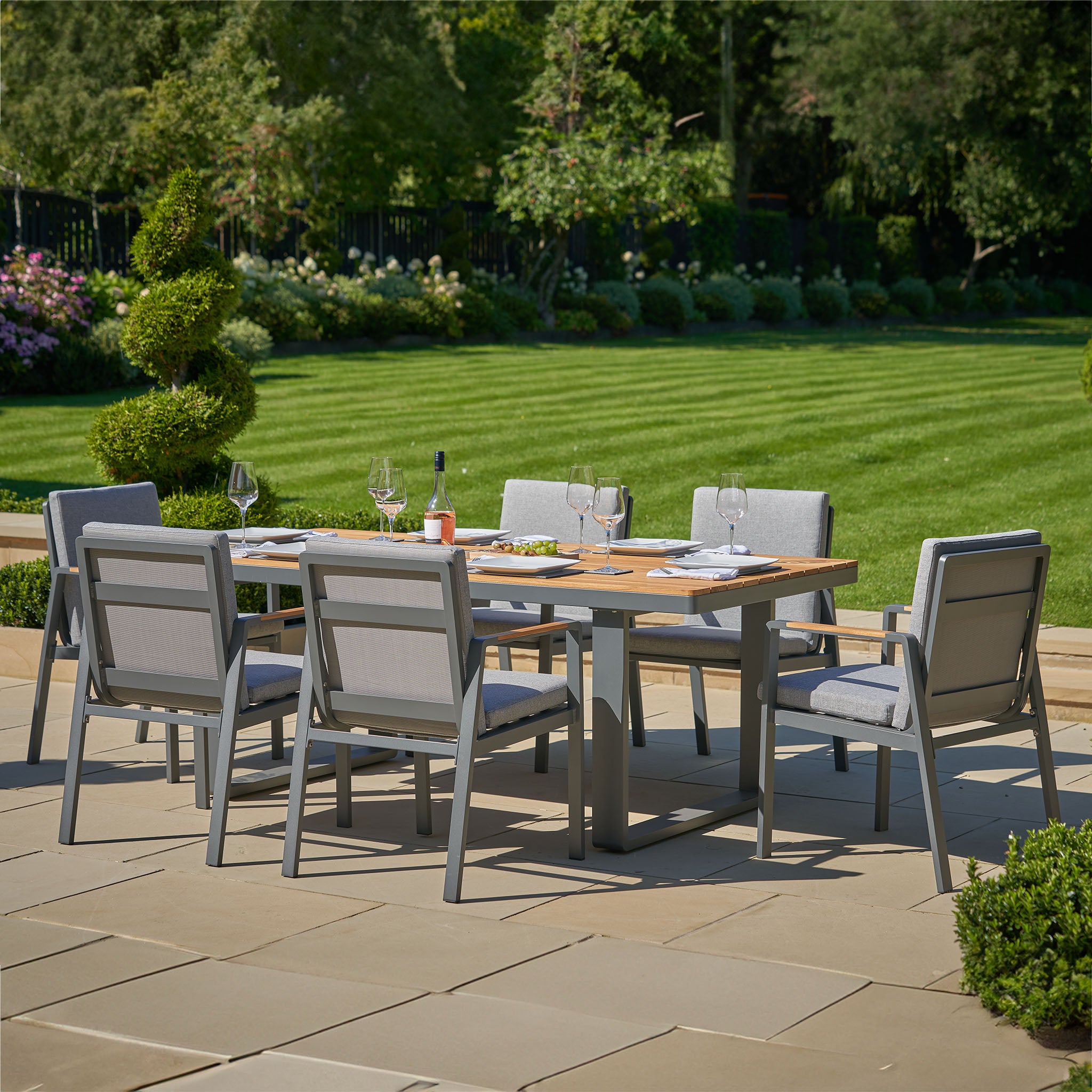 The Pacific Lifestyle Stockholm 6 Seat Rectangular Dining Set in Marl Grey, featuring weatherproof chairs, is ideally placed on a patio with a view of the manicured lawn and garden.
