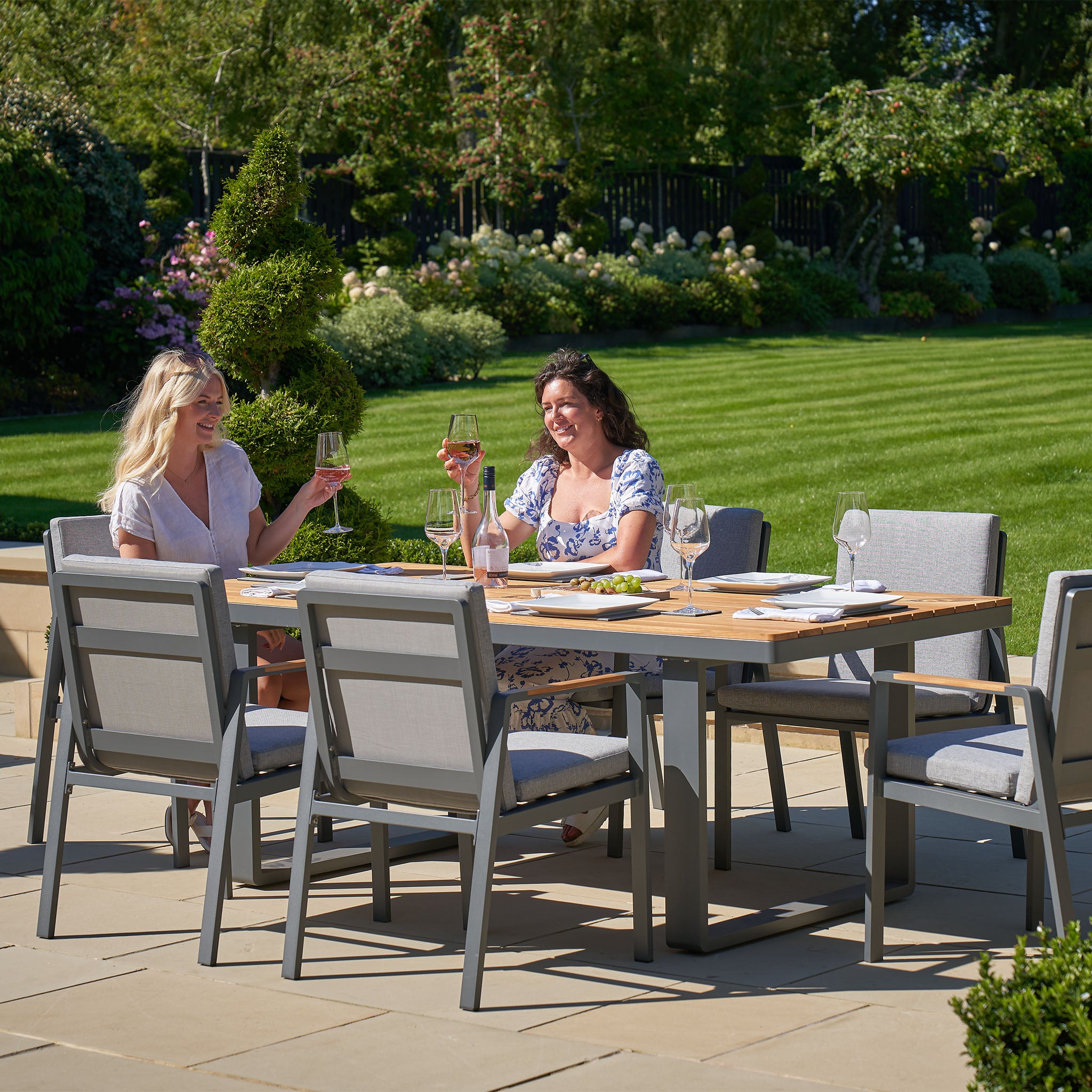 Two people sit at a patio table in a garden, toasting with wine glasses. They're surrounded by lush greenery and an elegant outdoor dining set featuring the weatherproof Pacific Lifestyle Stockholm 6 Seat Rectangular Dining Set in Marl Grey.