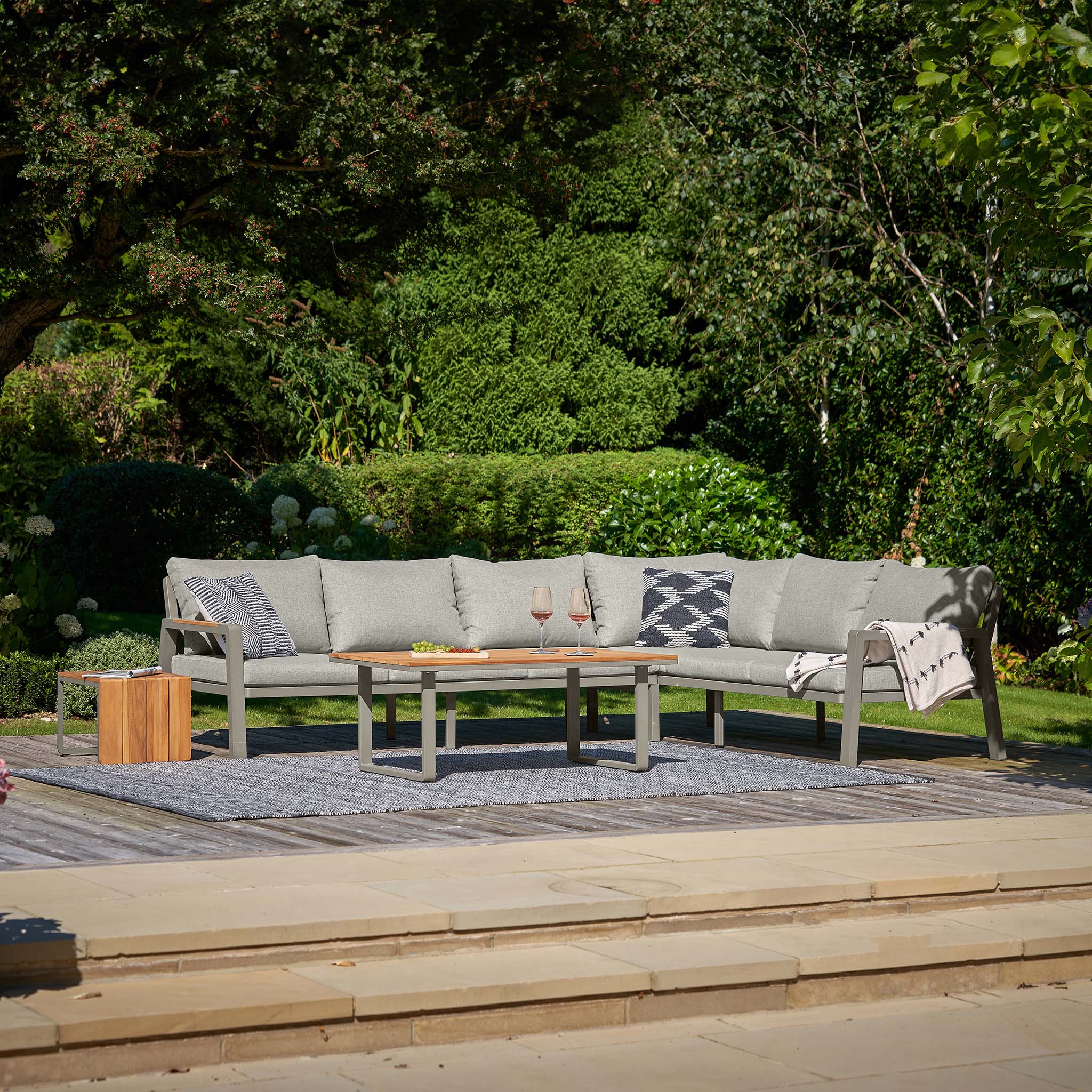 An outdoor patio showcases the Pacific Lifestyle - Stockholm Corner Set in Limestone, featuring an L-shaped sofa and a wooden coffee table with teak detailing. Two wine glasses sit on a rug, surrounded by lush greenery.