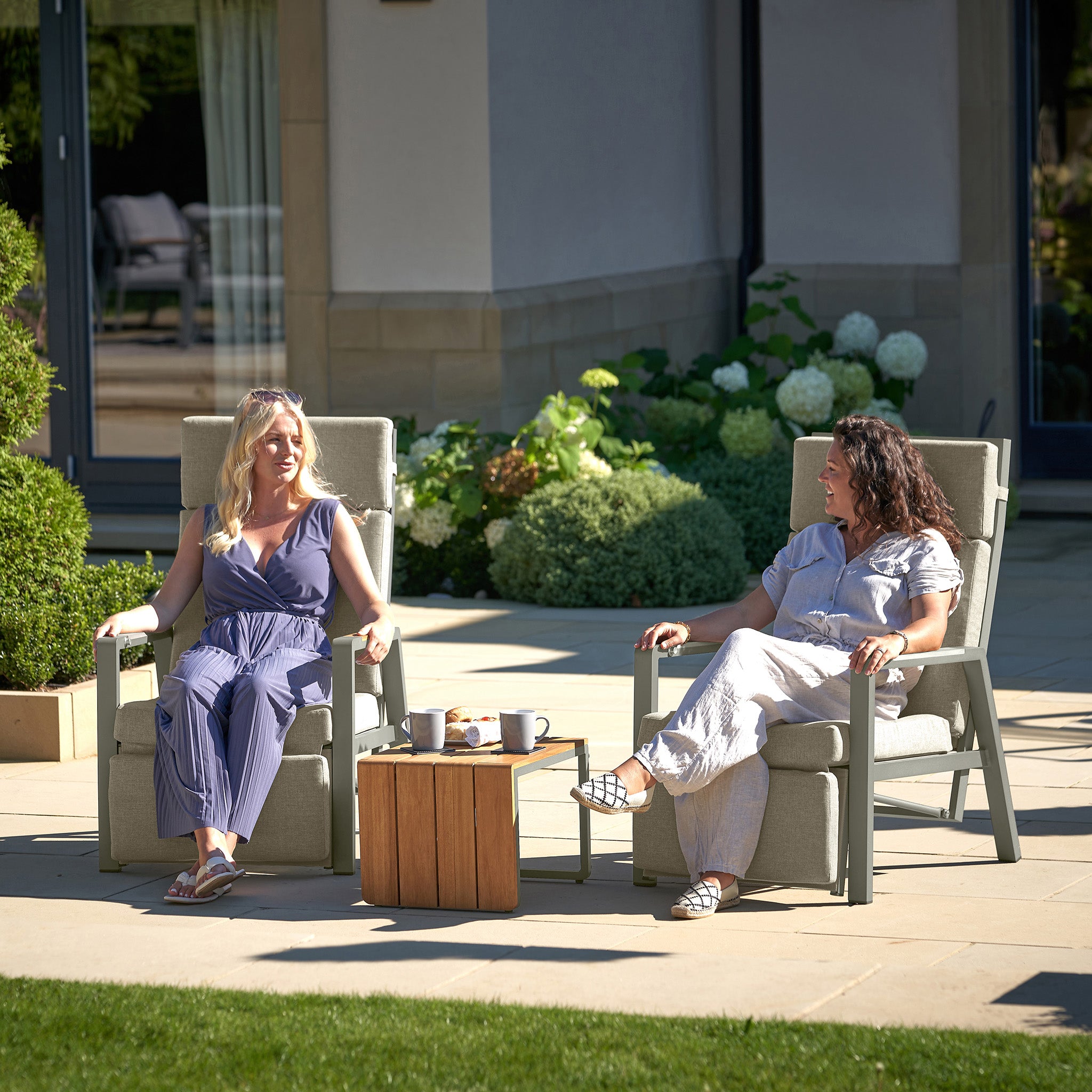 Two women sit on Pacific Lifestyle's Stockholm Lounge Reclining Set in Limestone, smiling and chatting with drinks on a small table between them. Surrounded by lush greenery, their garden lounging is enhanced with weatherproof cushions for comfort in any season.