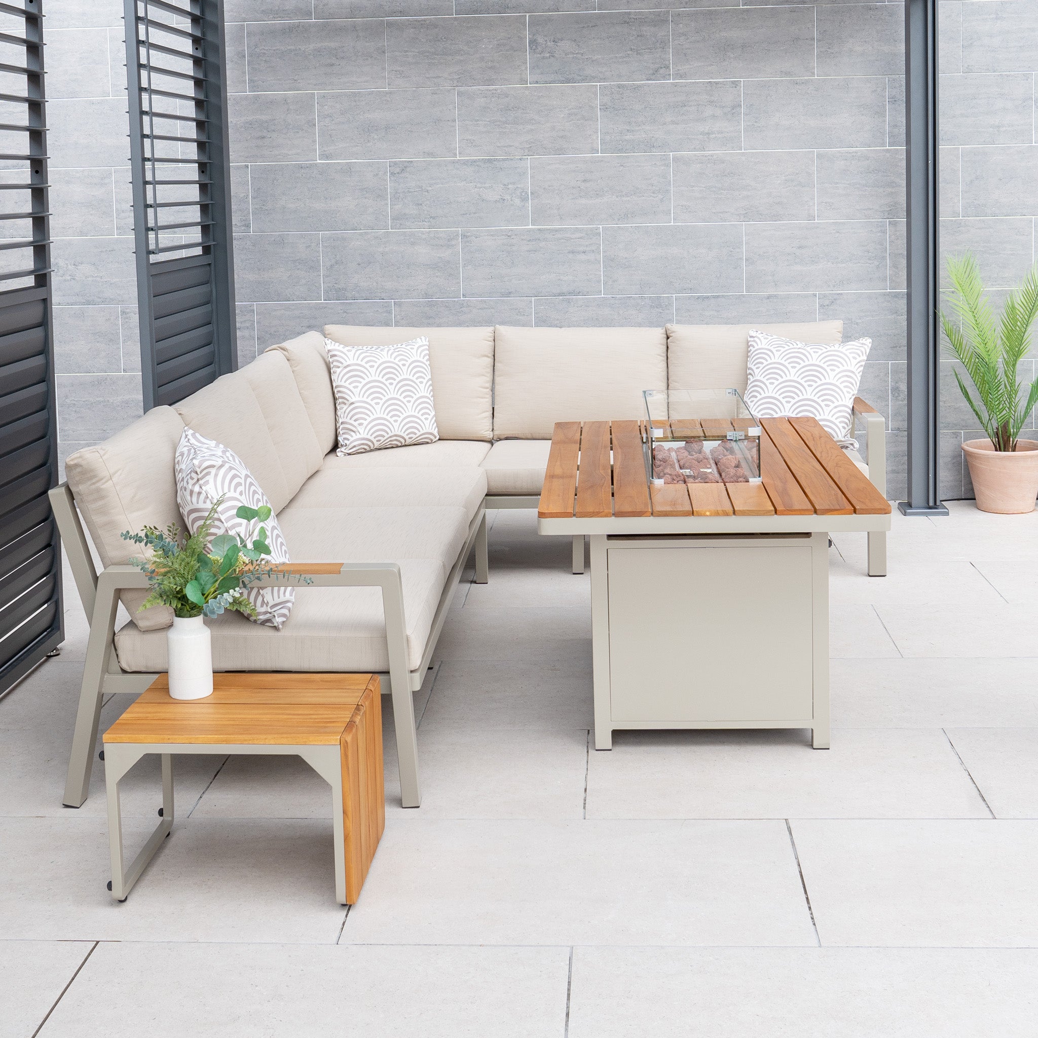Outdoor patio showcasing a beige sectional from the Pacific Lifestyle - Stockholm Corner Dining Set with Firepit Table in Limestone, complemented by a teak wood coffee table, small side table, and potted plant against a gray wall.