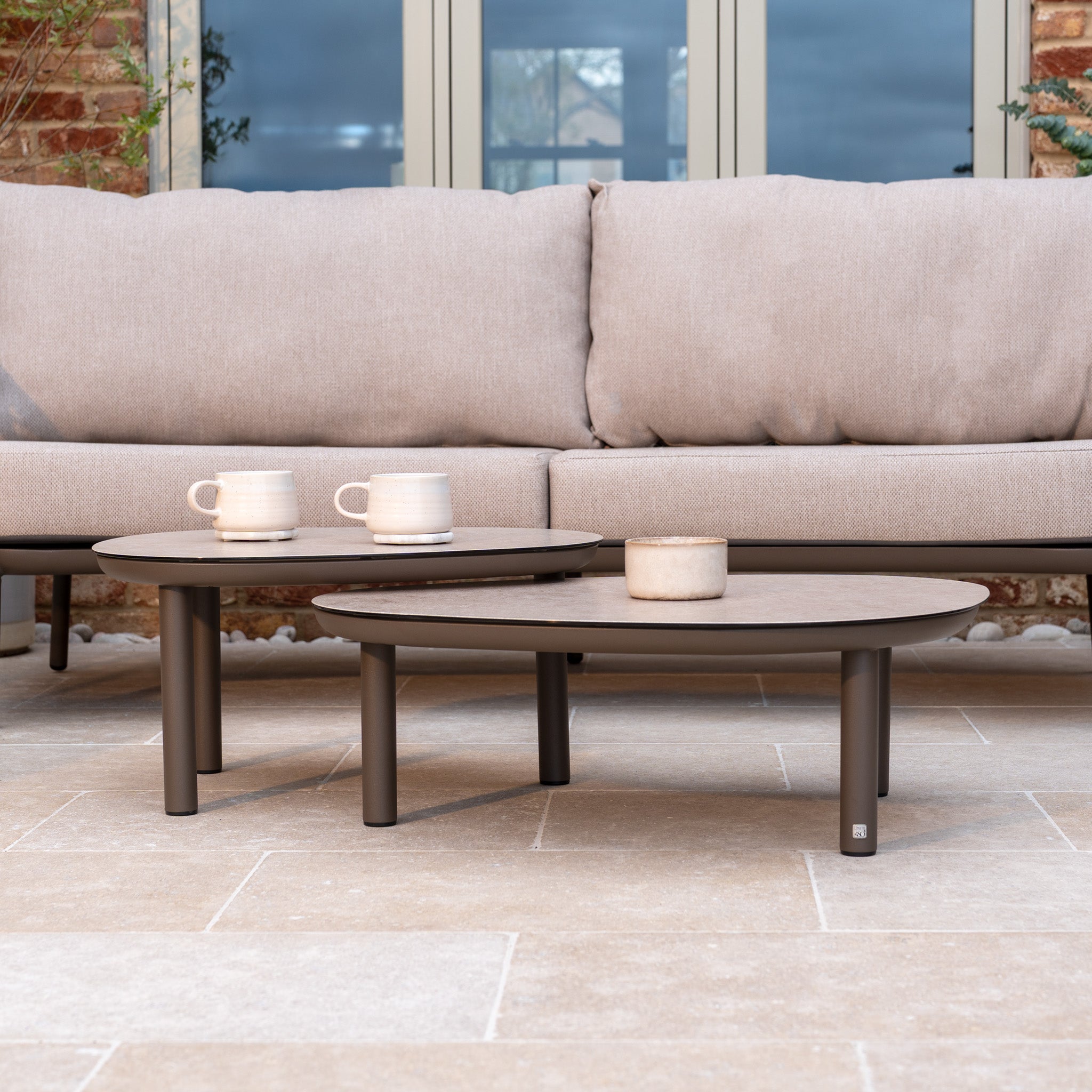 Two beige coffee tables with mugs and a small pot sit in front of the Sutera 3 Seat Sofa Set in Putty, featuring all-weather cushions for the perfect outdoor living space on your patio.