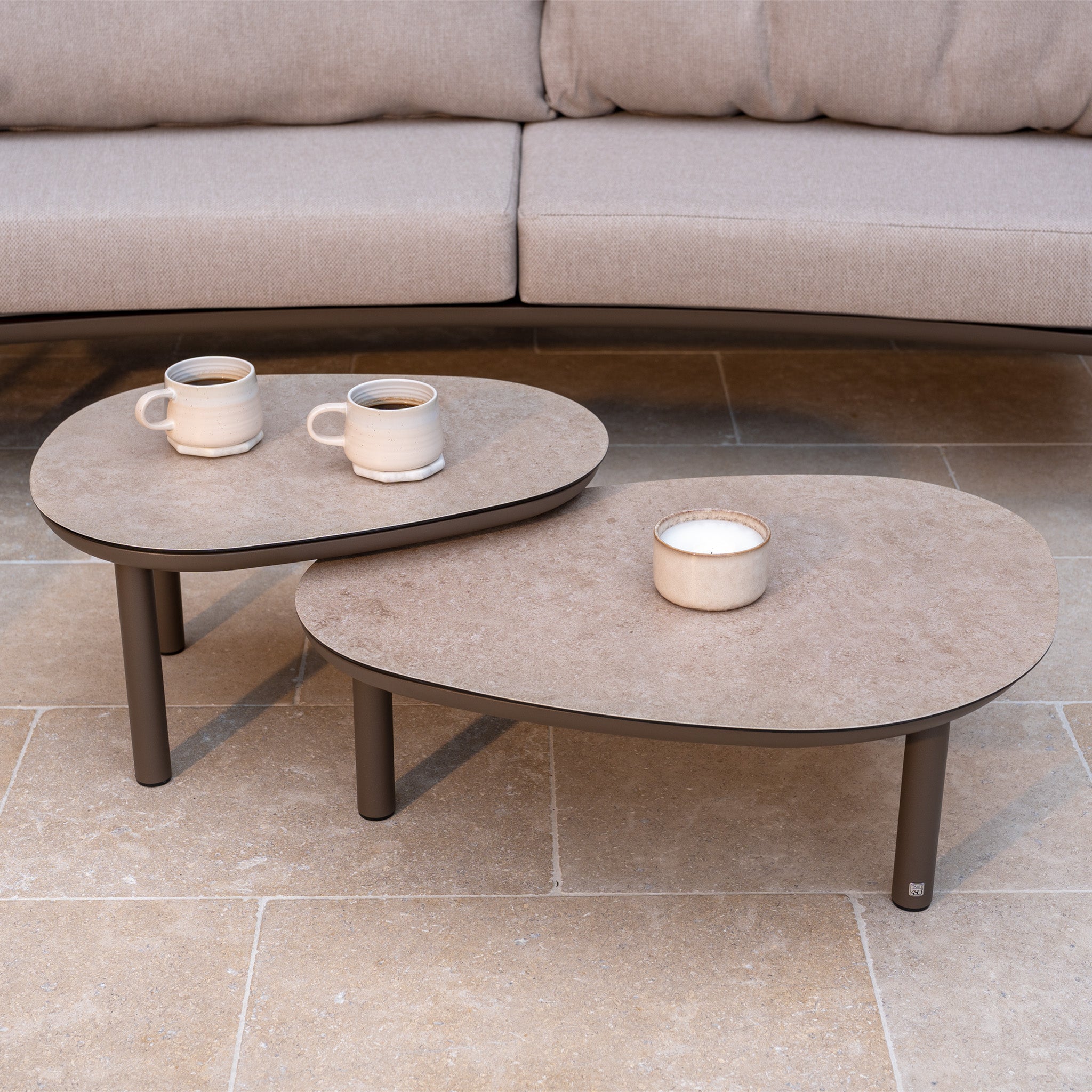 Two beige coffee tables with mugs and a candle are in front of the Sutera 3 Seat Sofa Set in Putty with all-weather cushions, perfect for stylish outdoor living on a tiled floor.