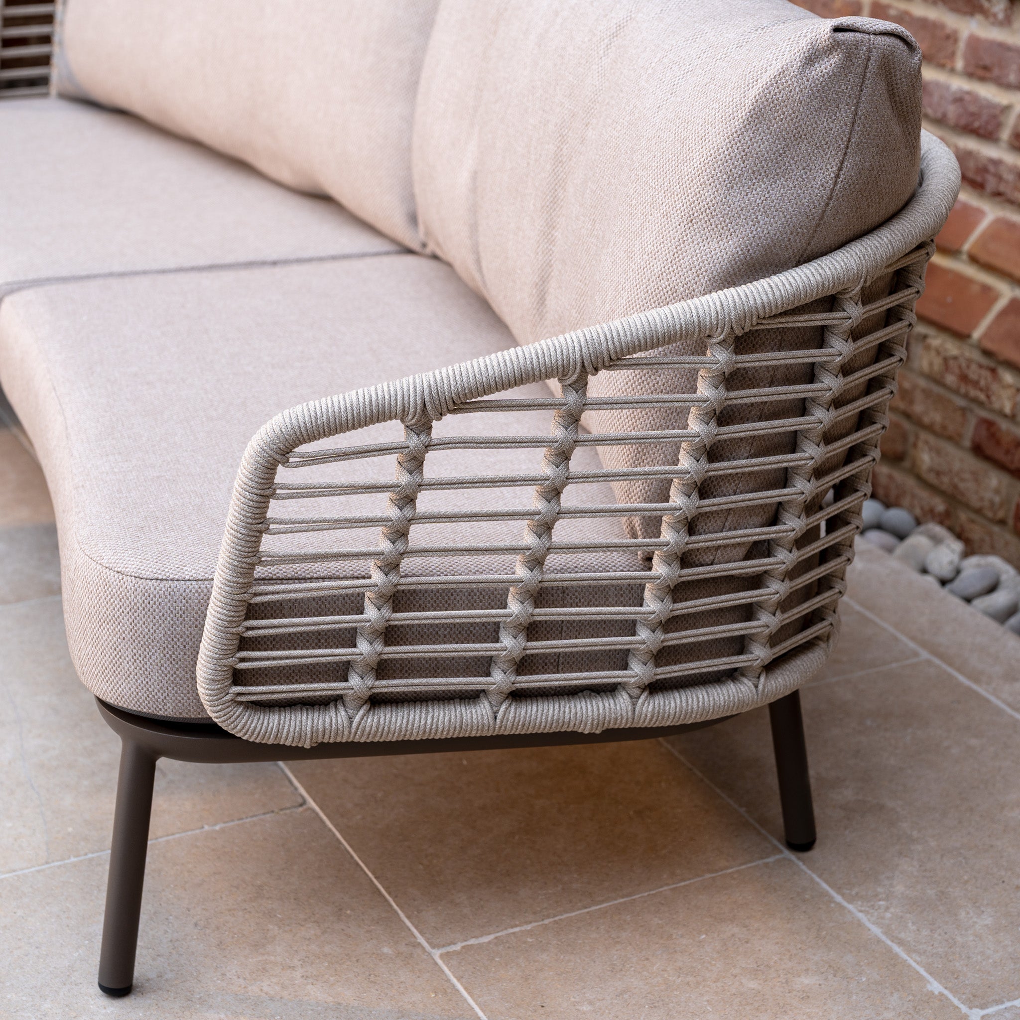 The Sutera 3 Seat Sofa Set in Putty features woven rope armrests and all-weather cushions, ideal for outdoor living on a tiled patio next to a brick wall.