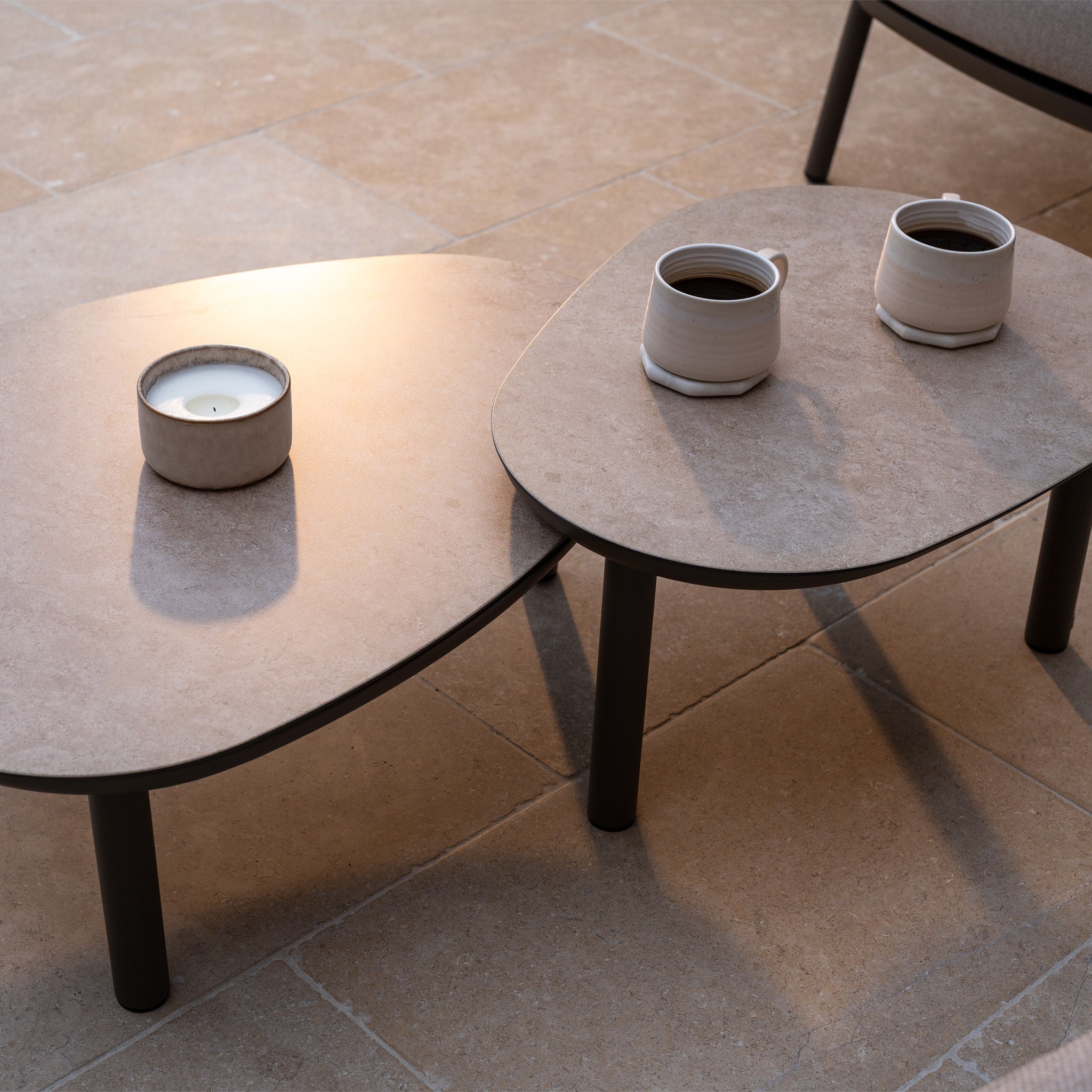 The Sutera 3 Seat Sofa Set in Putty, with all-weather cushions, is arranged beside two beige tables topped with a candle and coffee mugs, all set on a tiled floor bathed in soft natural light.