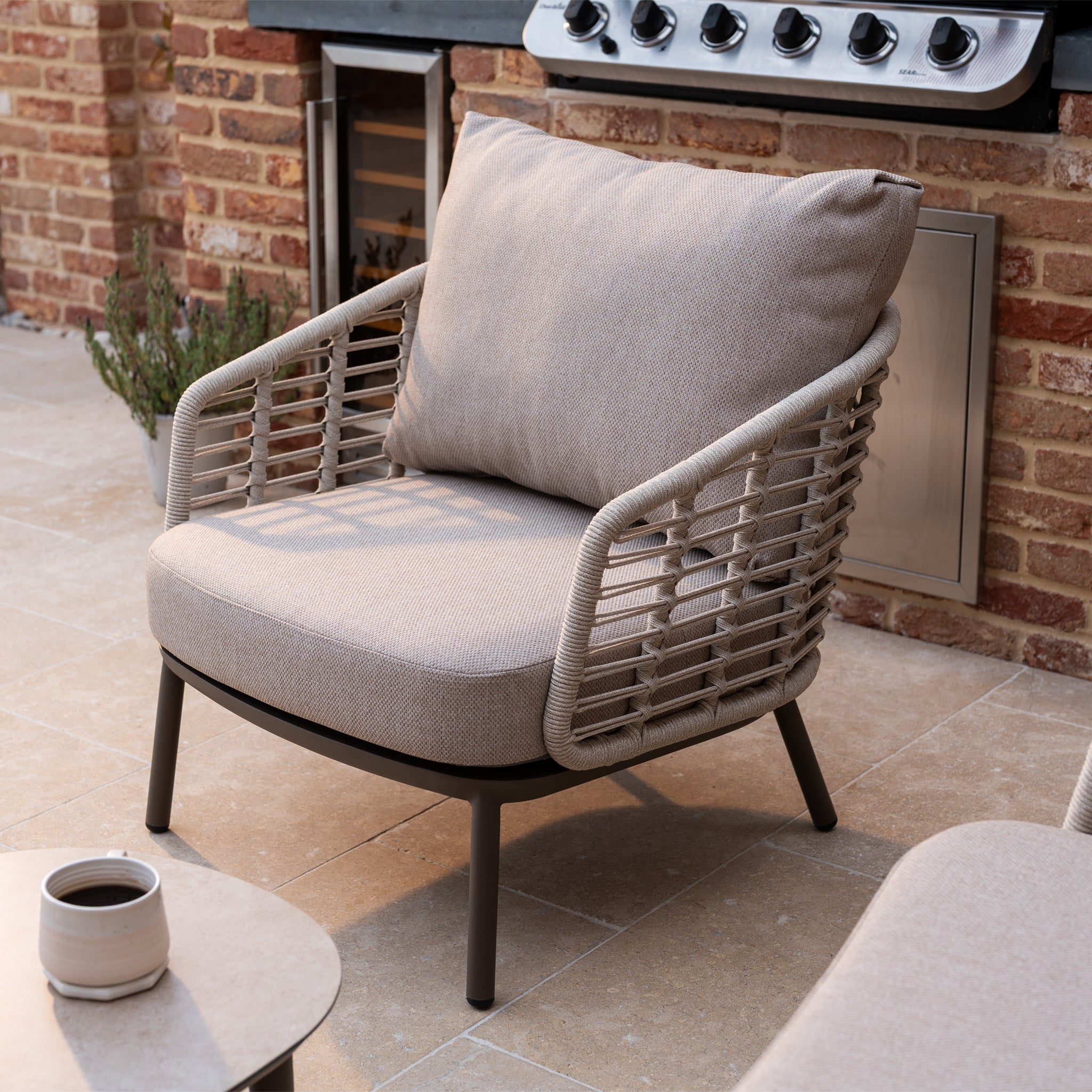 The Sutera 3 Seat Sofa Set in Putty features rope sides and all-weather cushions, ideal for outdoor living on your patio—shown beside a table with a cup and a grill in the background.