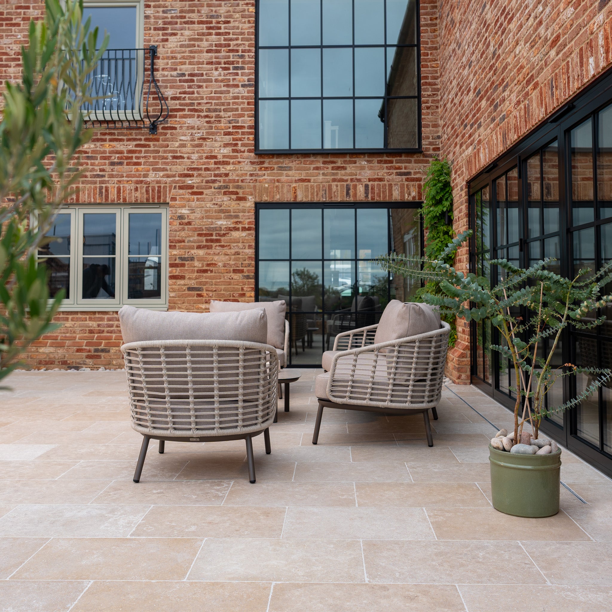 Enjoy outdoor living with the Sutera 3 Seat Sofa Set in Putty, featuring wicker design, all-weather cushions, and a sleek table—ideal for relaxing on your modern brick patio near large glass windows with potted plants.