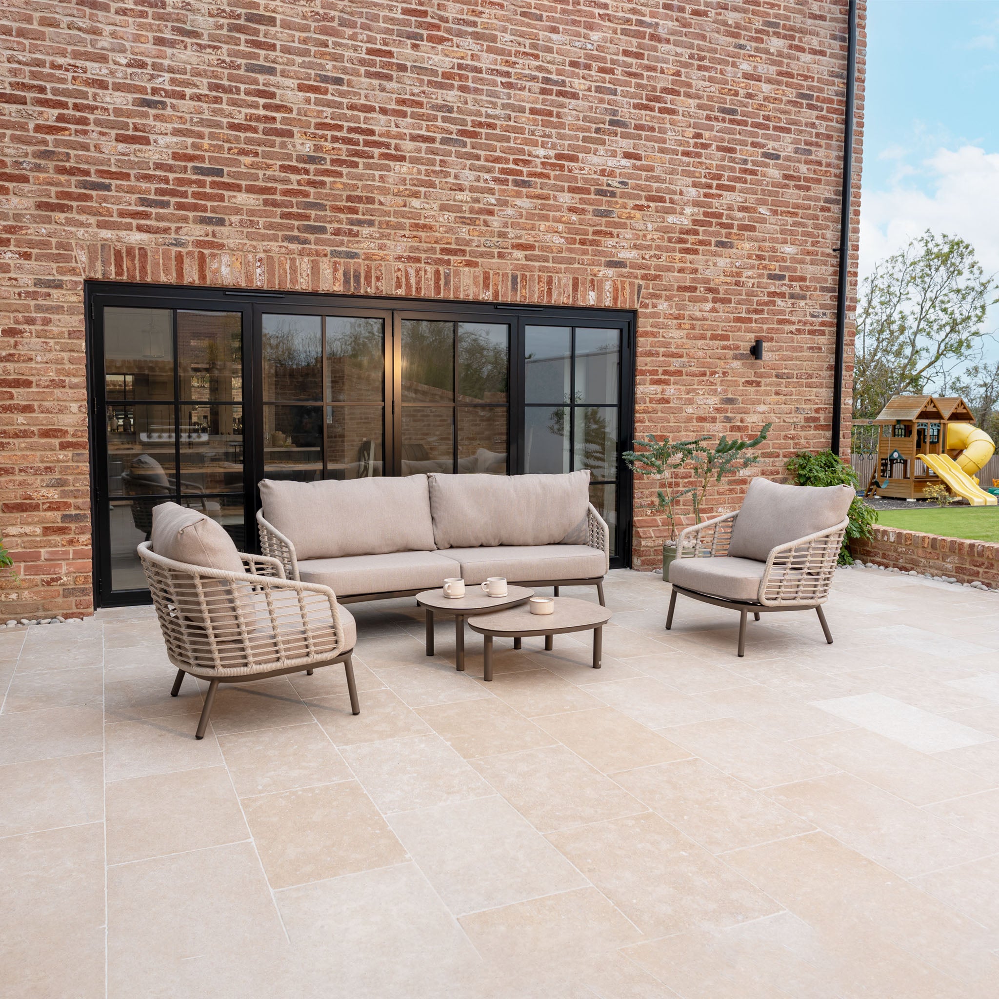 Outdoor patio with the Sutera 3 Seat Sofa Set in Putty, a matching coffee table, beige all-weather cushions, brick wall backdrop, and a playground set in the background.
