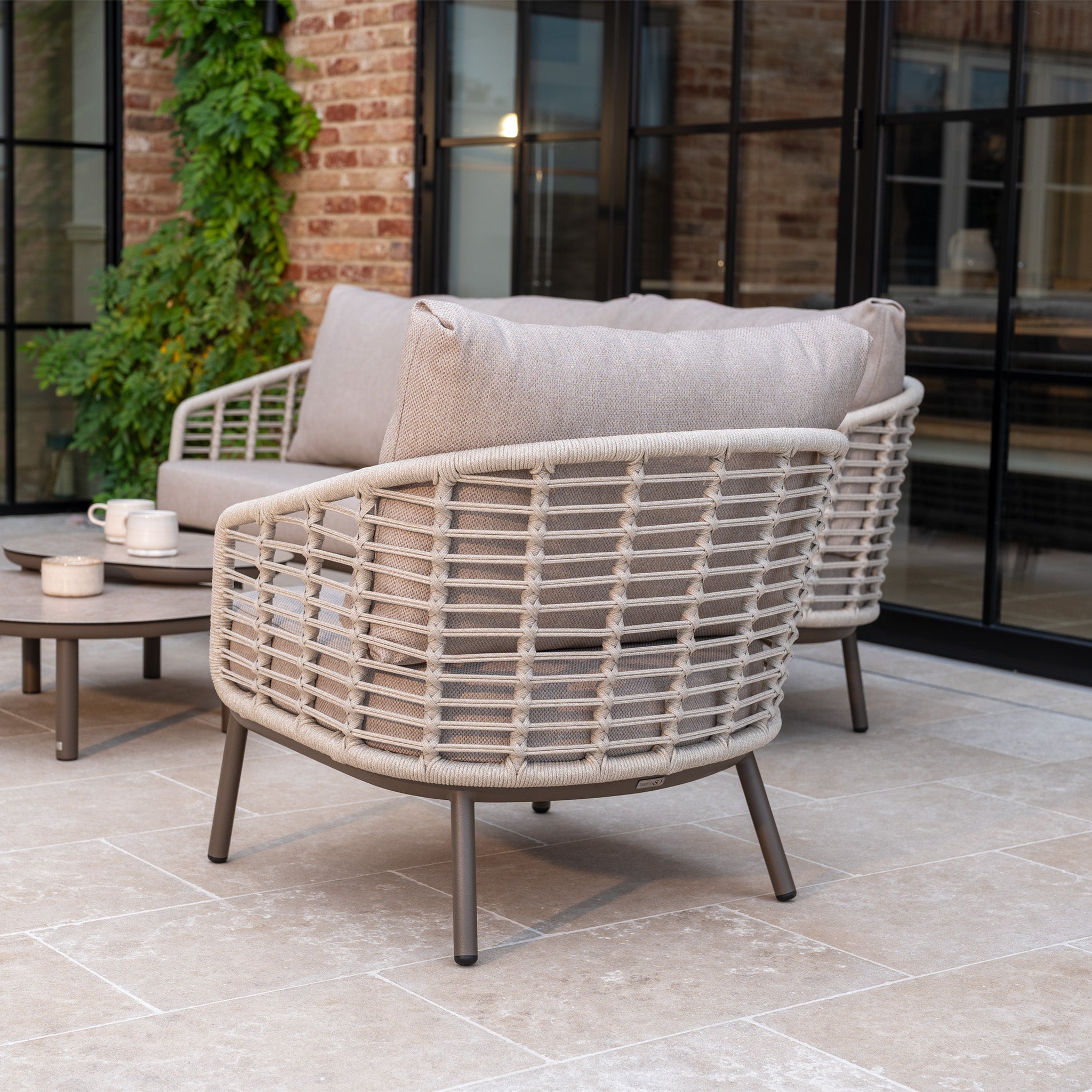 The Sutera 3 Seat Sofa Set in Putty creates the perfect outdoor living space on a tiled patio outside a brick building with large windows.