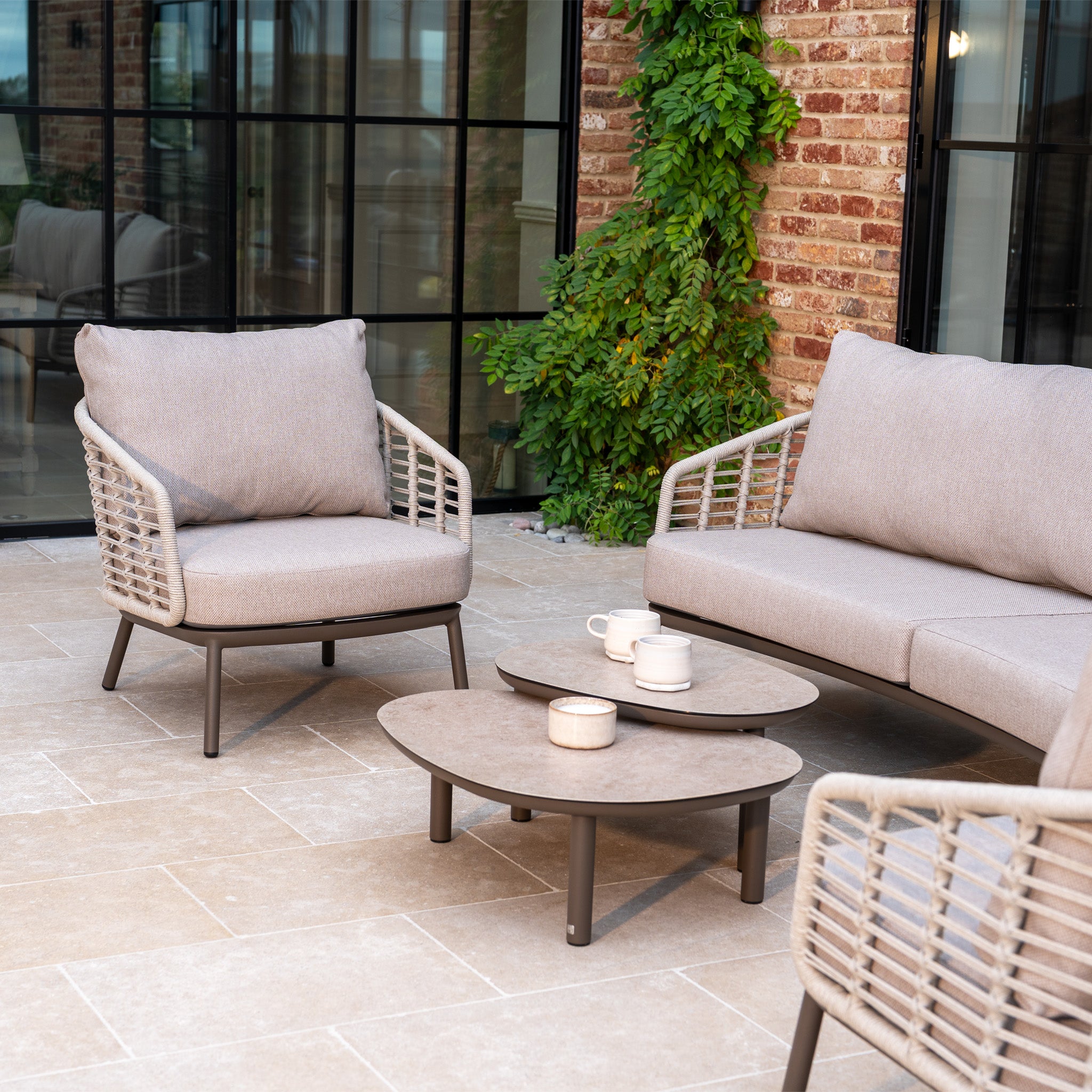 Modern patio seating with the Sutera 3 Seat Sofa Set in Putty, featuring cushioned chairs and a round coffee table with mugs and a candle.