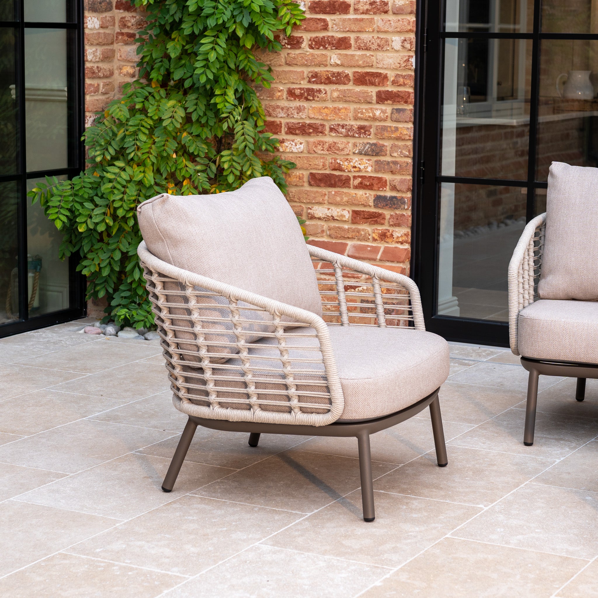 Beige woven outdoor chair with all-weather cushions, ideal for a tiled patio near a leafy brick wall. Pair it with the Sutera 3 Seat Sofa Set in Putty for a stylish, complete outdoor living setup.