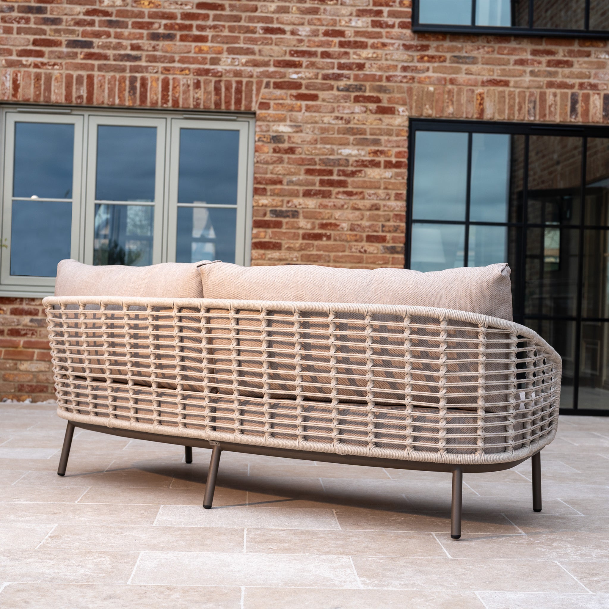 The Sutera 3 Seat Sofa Set in Putty with all-weather cushions is shown on a tiled patio, positioned in front of a brick house featuring large windows.