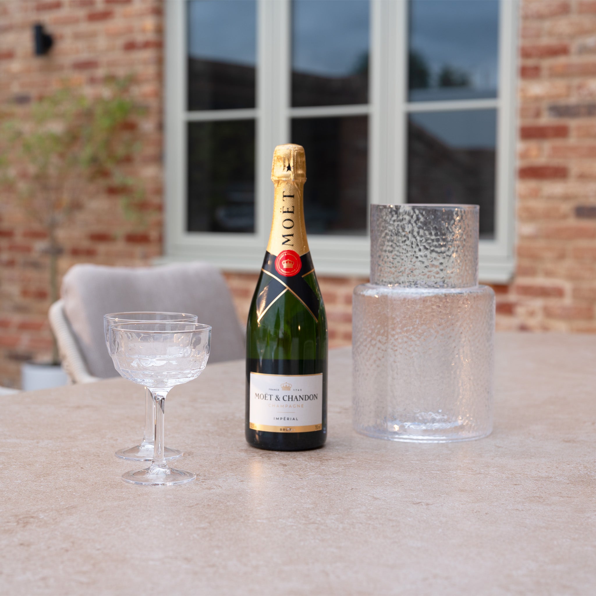 A bottle of Moët & Chandon champagne with two glasses and an ice bucket on a patio table, perfectly complementing the Sutera 6 Seat Curved Rectangular Dining Set in Putty for stylish outdoor dining.