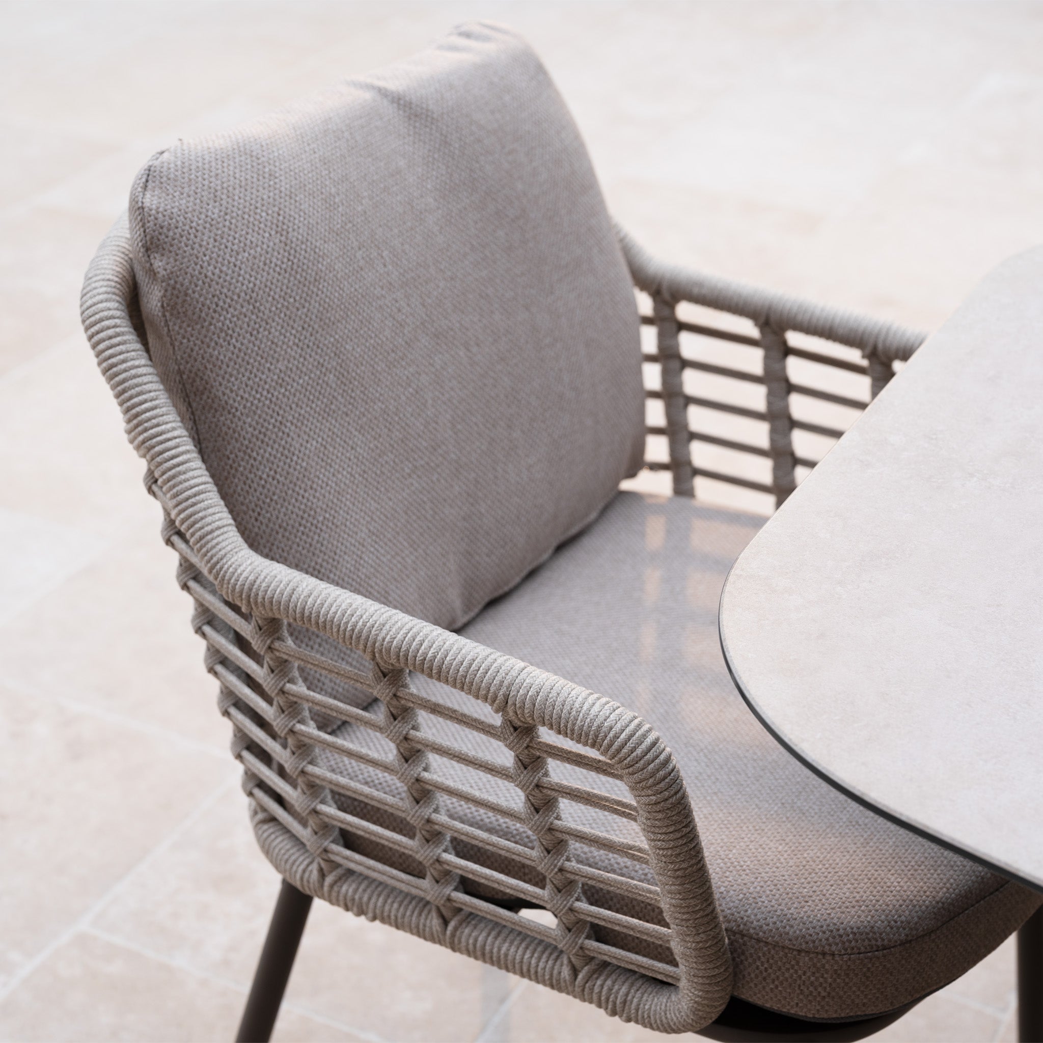 The Sutera 6 Seat Curved Rectangular Dining Set in Putty features a woven rope armchair with grey cushions beside a light table on tiled flooring. All-weather fabric ensures lasting comfort for outdoor dining.