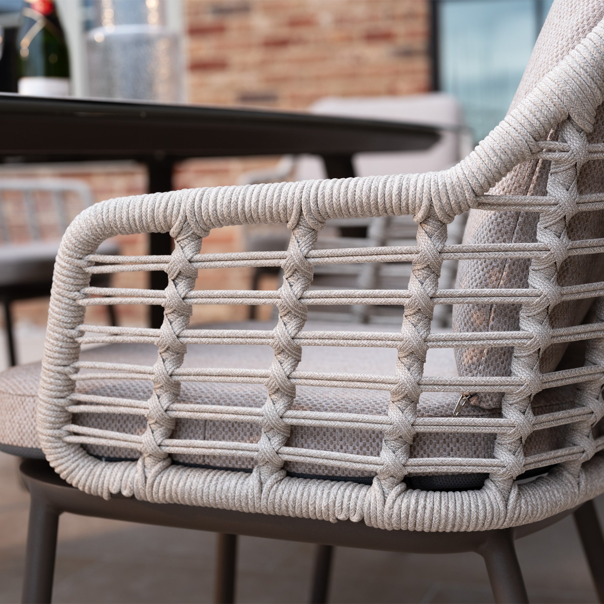 Close-up of a woven rope armrest on the Sutera 6 Seat Curved Rectangular Dining Set in Putty, shown with table and brick wall in the background. Crafted with all-weather fabric, it’s ideal for any outdoor occasion.