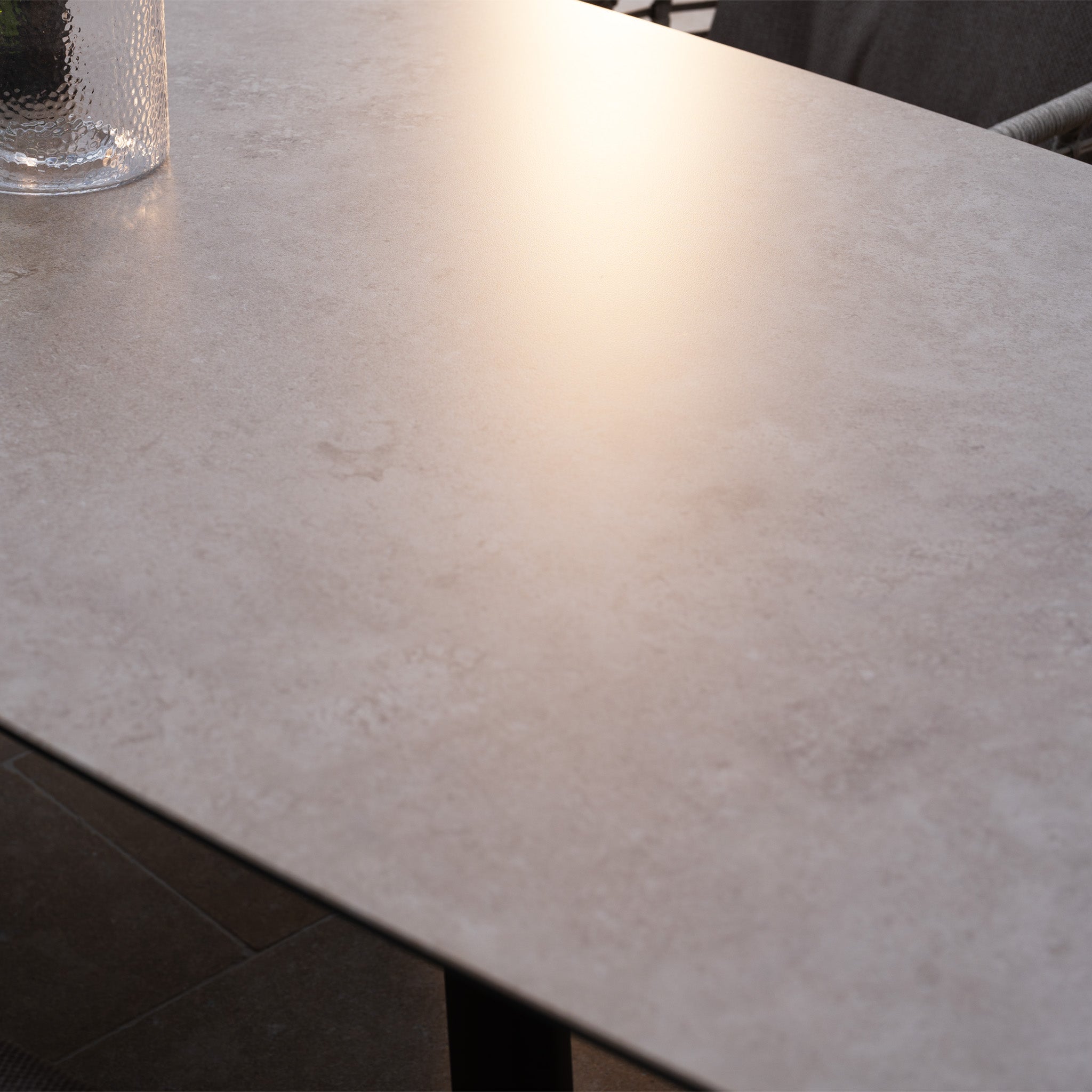 Sunlight reflects off the smooth, light tabletop of the Sutera 6 Seat Curved Rectangular Dining Set in Putty, with a textured glass vase nearby. Featuring all-weather fabric, it’s perfect for comfortable outdoor gatherings.