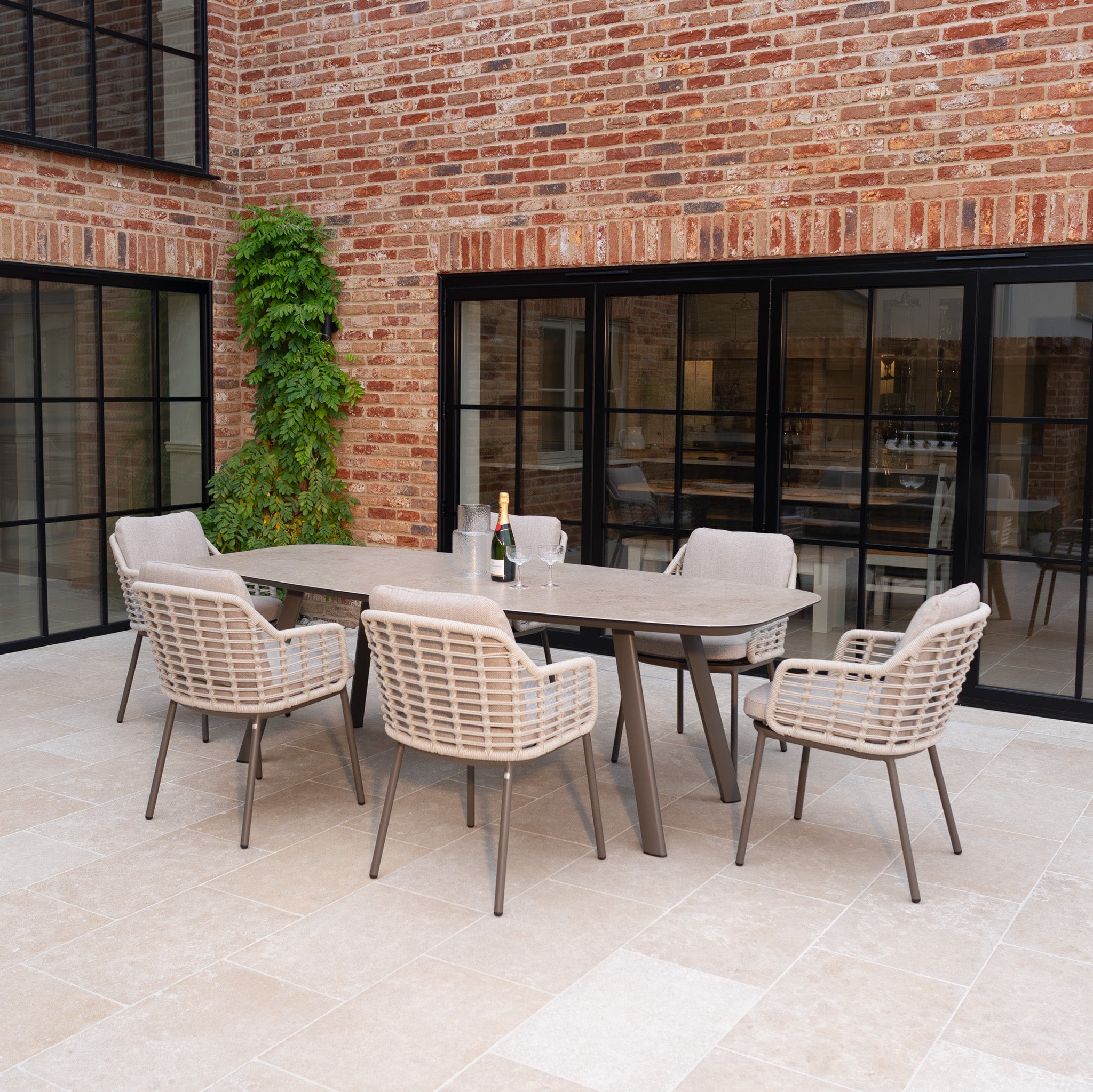The Sutera 6 Seat Curved Rectangular Dining Set in Putty features all-weather fabric chairs and a sleek table—ideal for stylish outdoor dining on a tiled patio with brick walls and a bottle of wine.