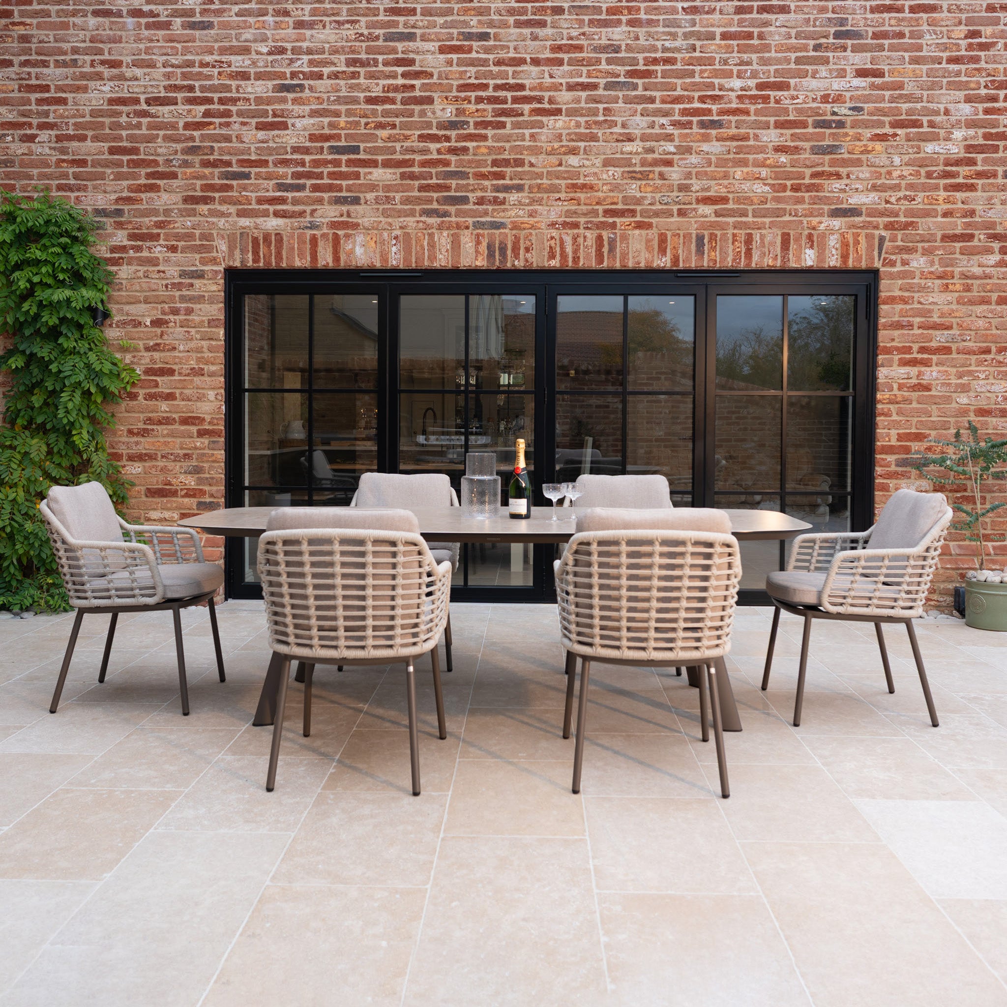 The Sutera 6 Seat Curved Rectangular Dining Set in Putty, crafted with all-weather fabric, is showcased on a patio with glasses and a bottle, set against a brick wall.