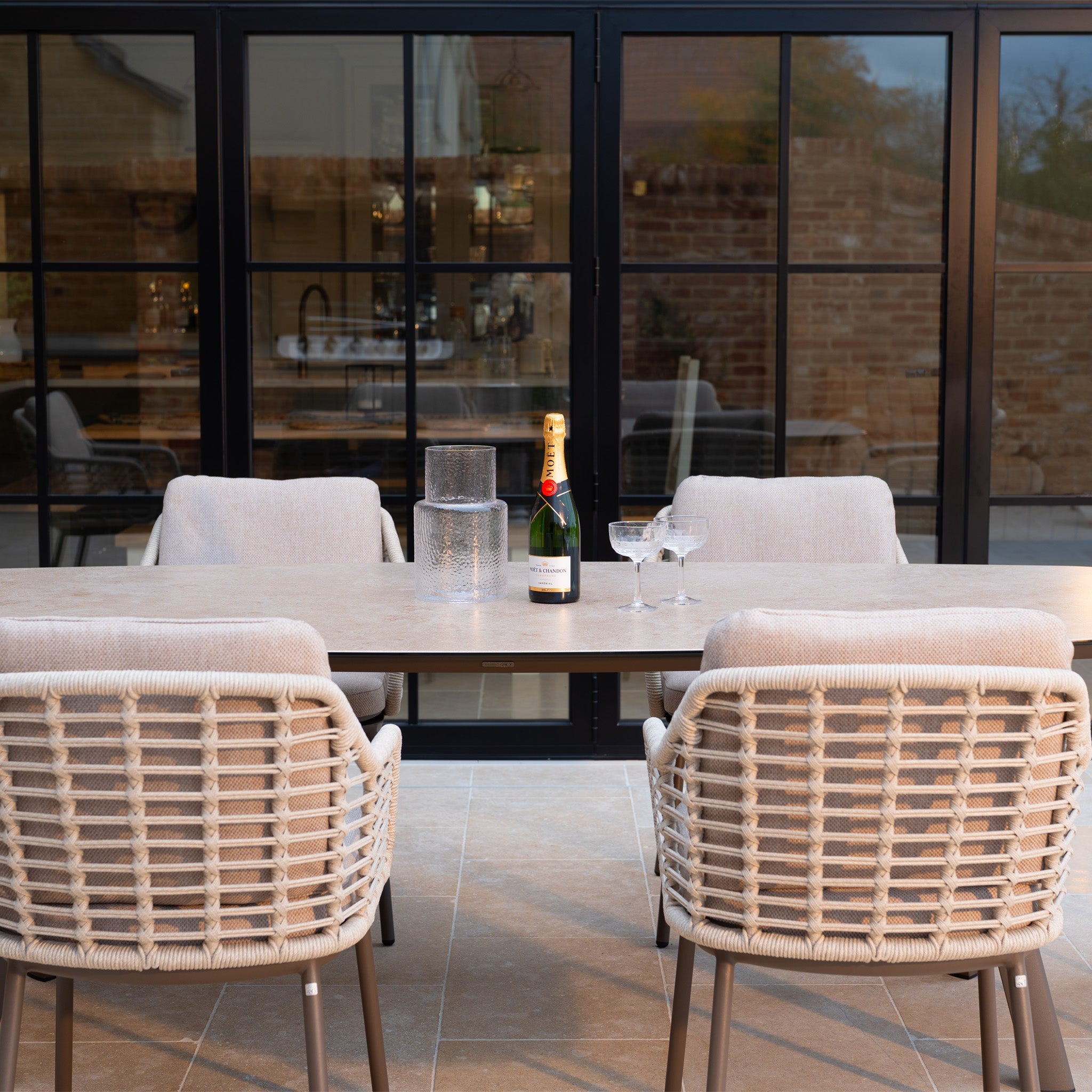 The Sutera 6 Seat Curved Rectangular Dining Set in Putty features a table with six all-weather chairs, plus a bottle of champagne, two glasses, and a water pitcher—ideal for stylish and relaxed outdoor dining.
