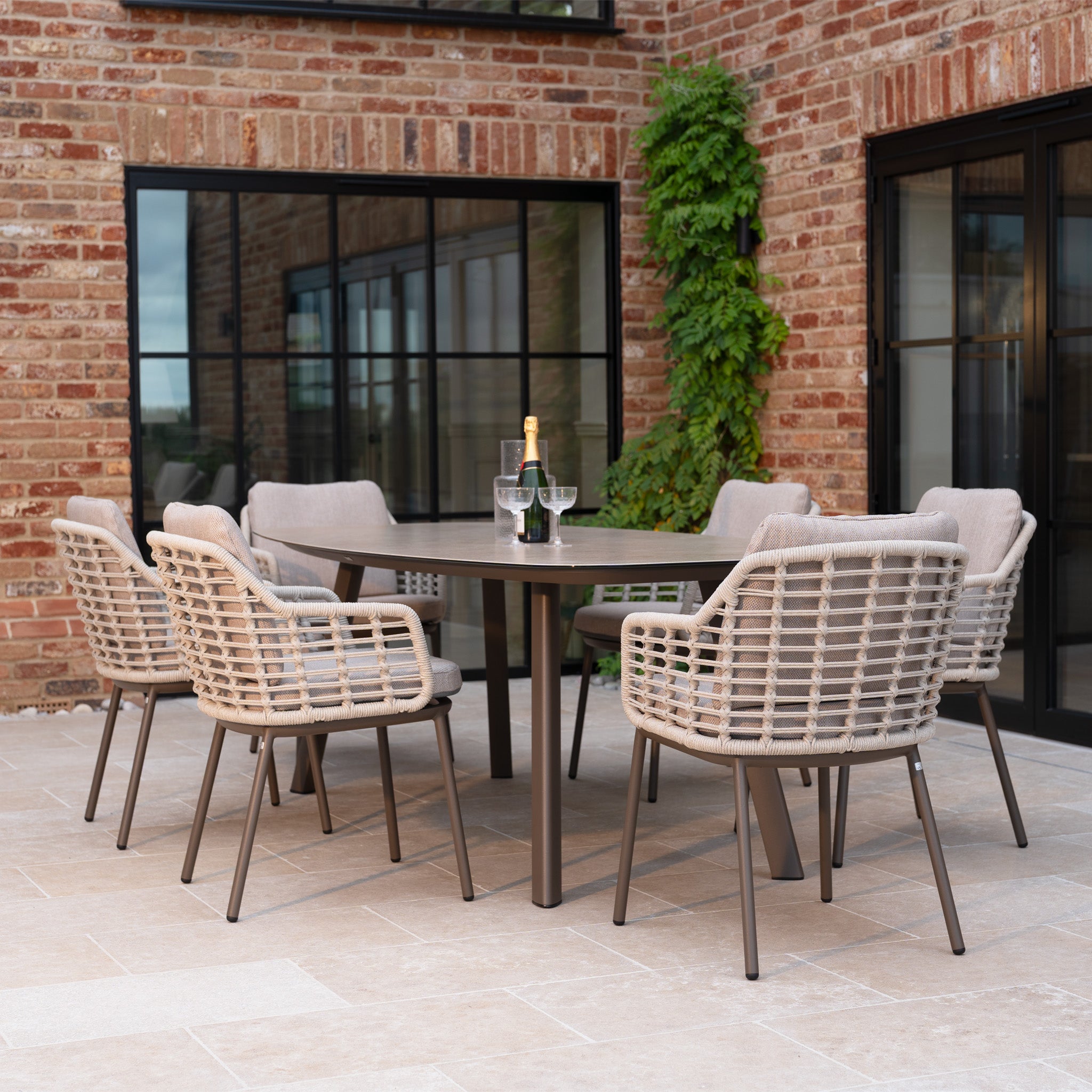 The Sutera 6 Seat Curved Rectangular Dining Set in Putty includes six wicker chairs with all-weather cushions and a rectangular table—ideal for outdoor meals with champagne and glasses.