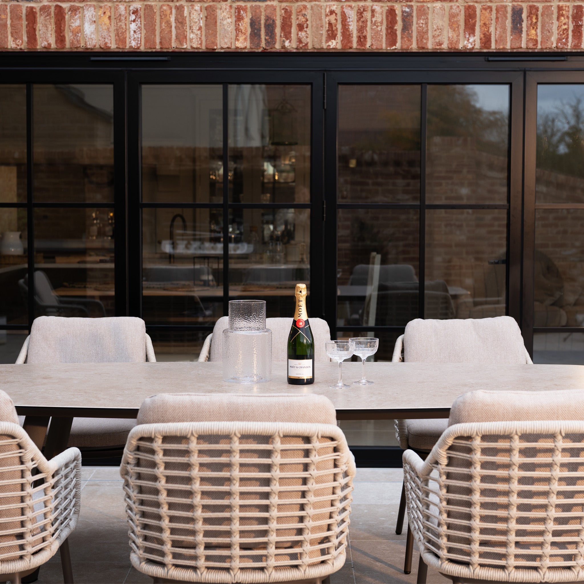 The Sutera 8 Seat Curved Rectangular Dining Set in Putty, complete with six chairs, a table, champagne bottle, and glasses, sits beautifully against a charming brick wall backdrop.