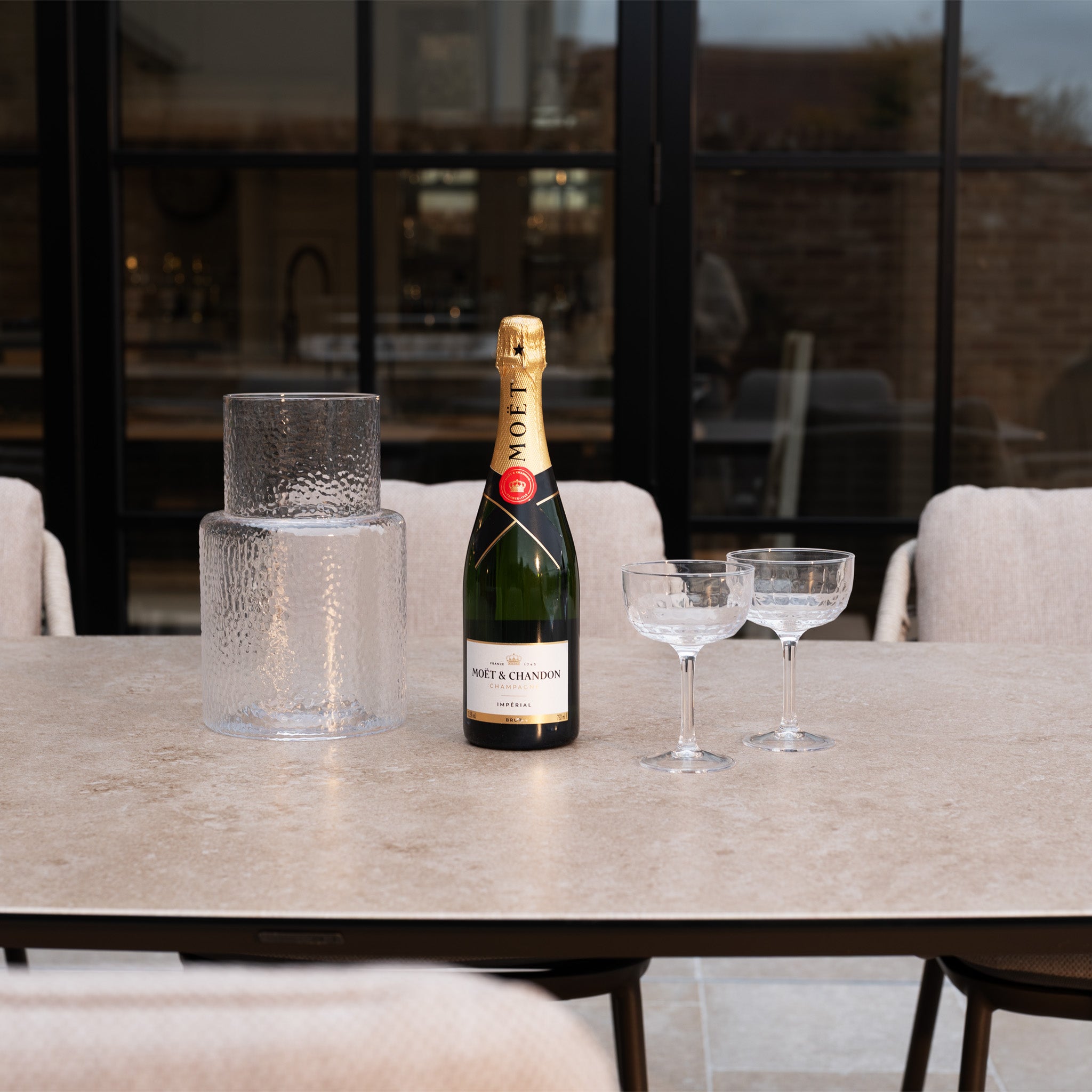A bottle of Moët & Chandon and two glasses sit beside a textured glass vase, all elegantly displayed on the Sutera 8 Seat Curved Rectangular Dining Set in Putty.