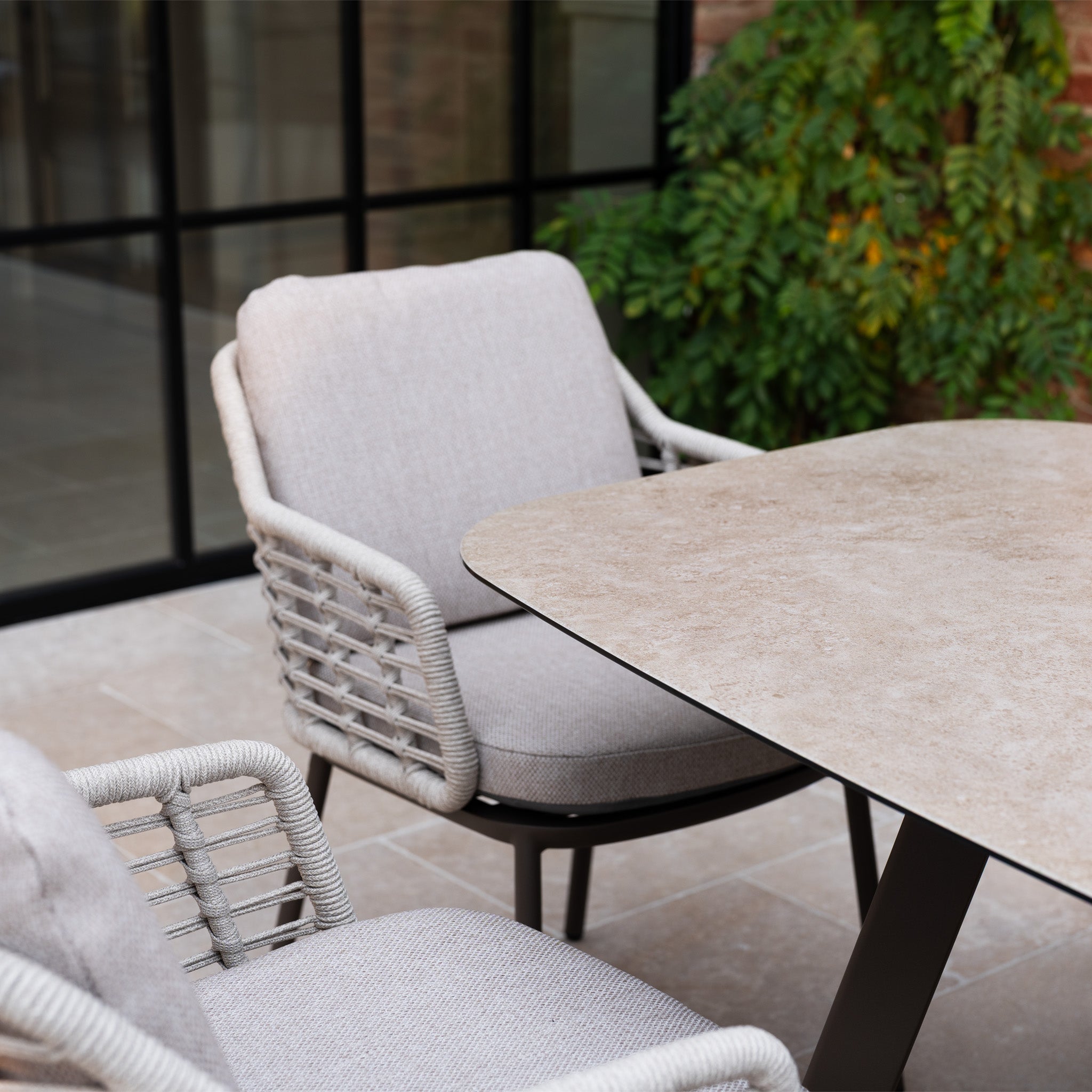 The Sutera 8 Seat Curved Rectangular Dining Set in Putty adds an inviting touch to your patio, featuring ample seating and contemporary style amidst natural greenery.