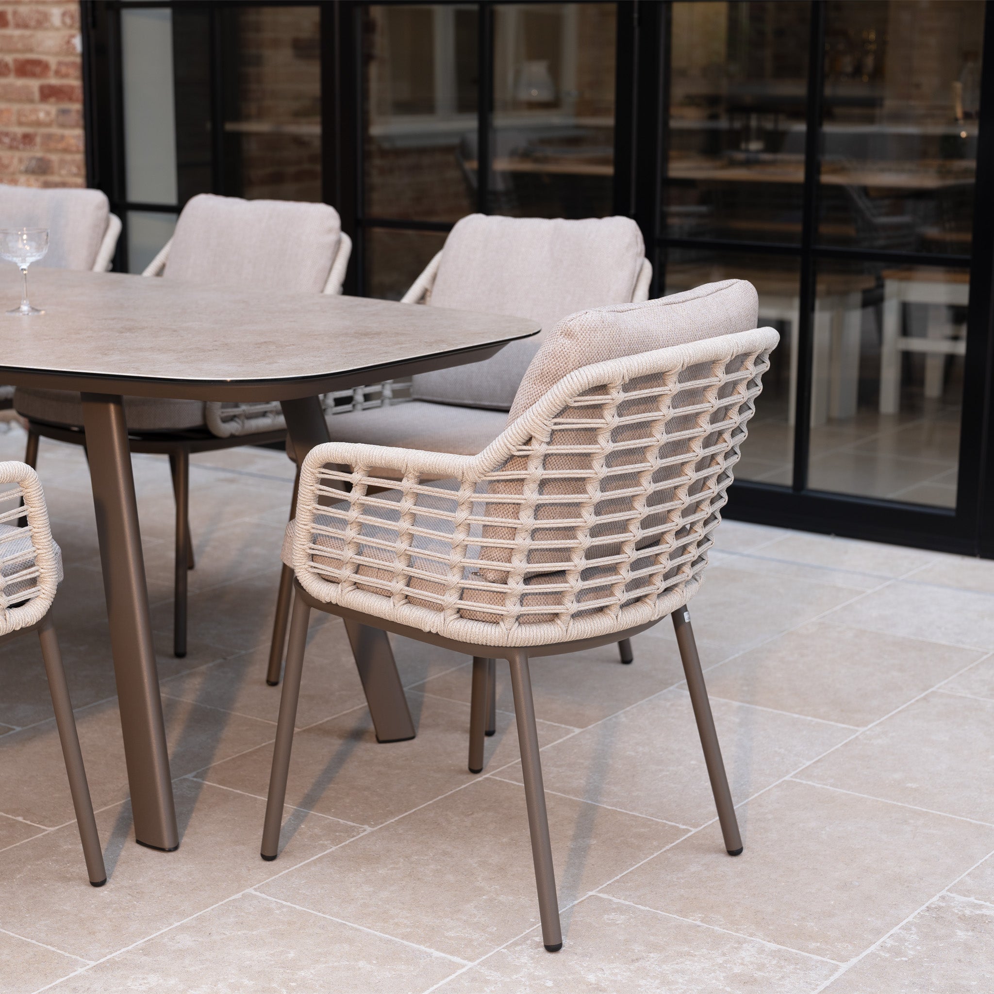 The Sutera 8 Seat Curved Rectangular Dining Set in Putty with cushioned chairs is arranged on a stone-tiled floor in a glass-enclosed patio.