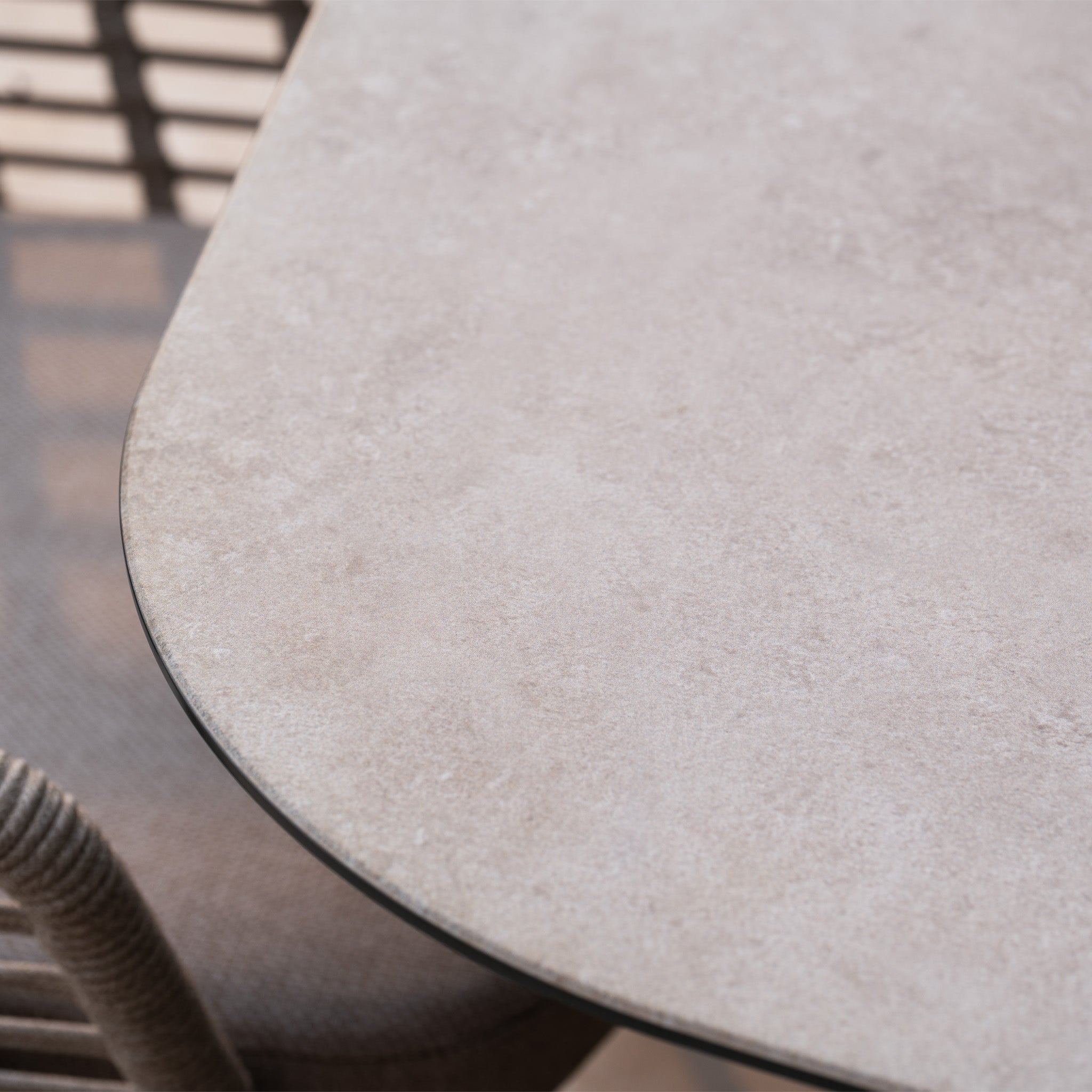 Close-up of the Sutera 8 Seat Curved Rectangular Dining Set in Putty, featuring a beige stone tabletop with a metal edge beside a woven chair—ideal for chic outdoor garden setups.