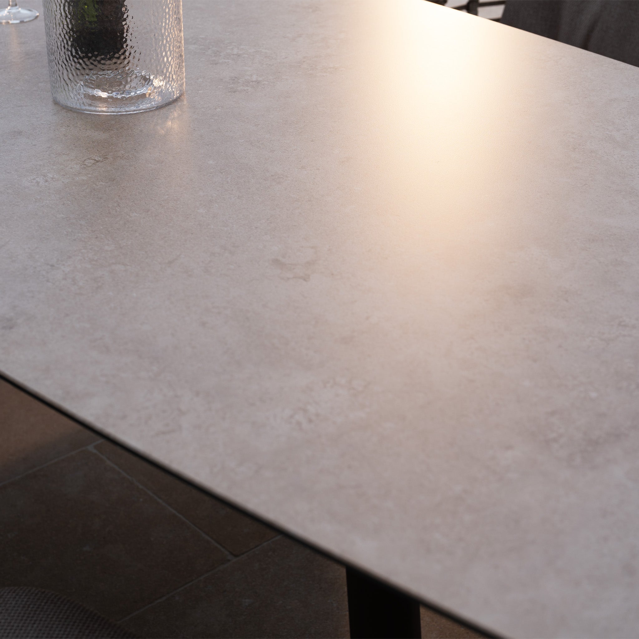 A glass of water rests on the sunlit stone table of the Sutera 8 Seat Curved Rectangular Dining Set in Putty, a soft reflection shimmering on its surface.