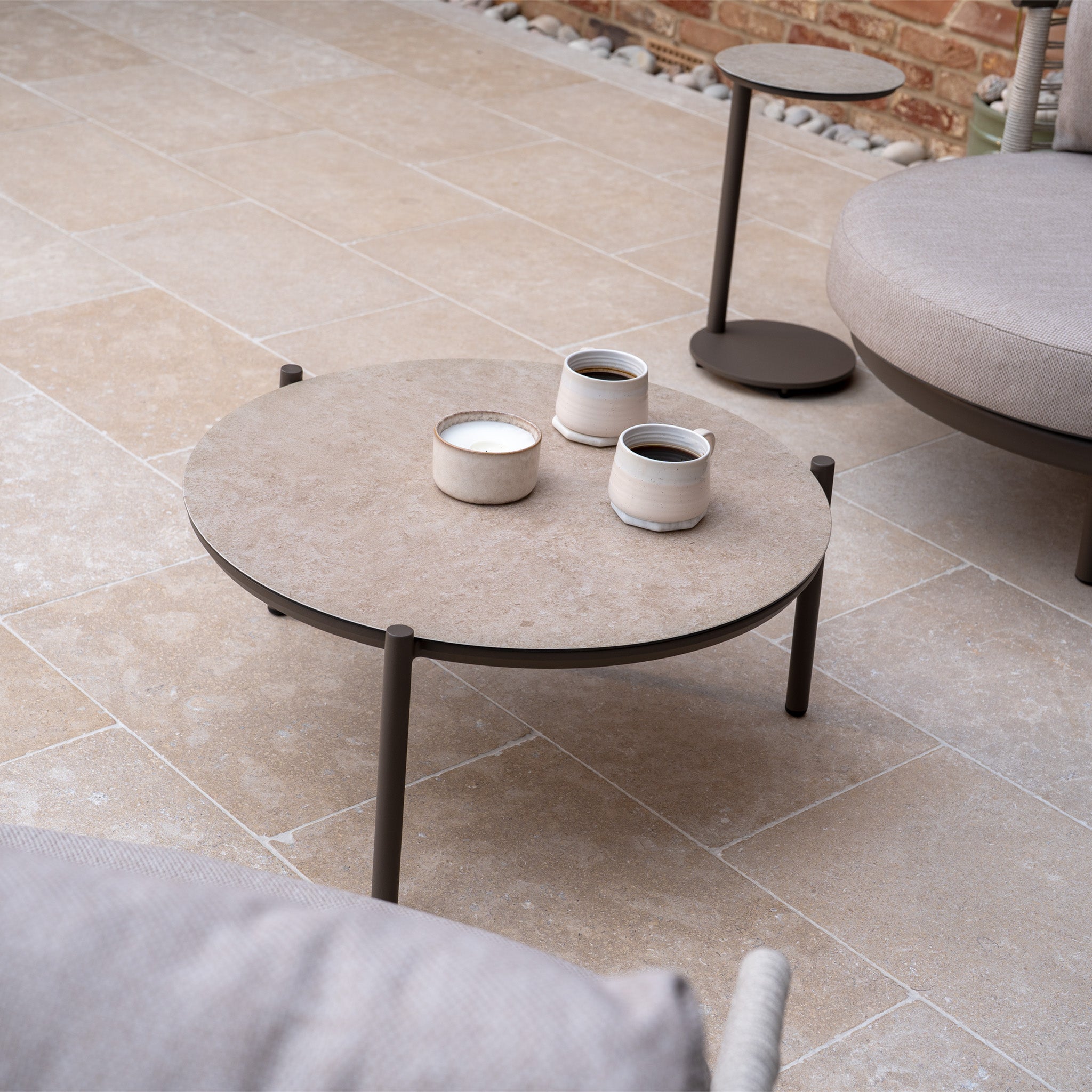 Round coffee table with three cups on a tiled patio, surrounded by the Sutera Curved Chaise Sofa Set in Putty, which features woven rope accents and plush all-weather cushions, set against a brick wall.