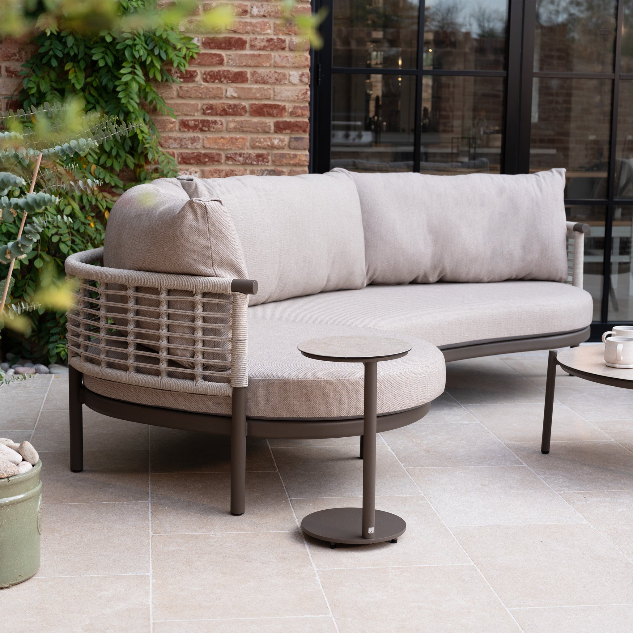 The Sutera Curved Chaise Sofa Set in Putty features all-weather cushions and woven rope accents, arranged with side tables on a tiled patio near glass doors and a brick wall.