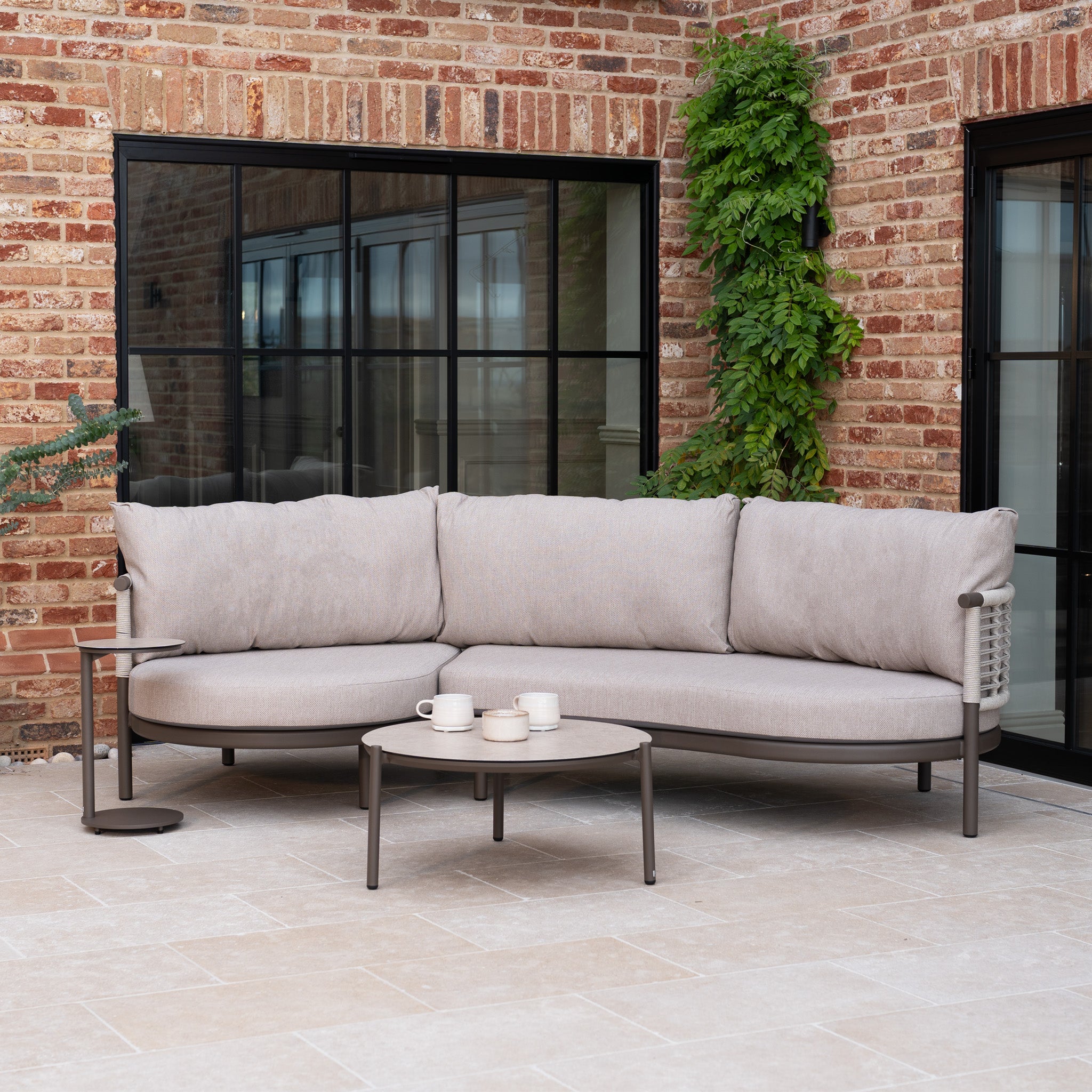 Sutera Curved Chaise Sofa Set in Putty, featuring all-weather cushions and woven rope accents, paired with a coffee table and side table on a tiled patio near a brick wall and greenery.