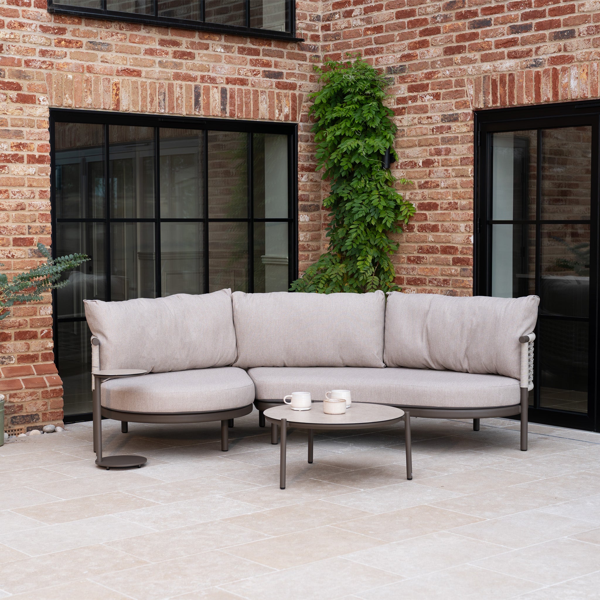 Modern outdoor patio with the Sutera Curved Chaise Sofa Set in Putty, featuring all-weather cushions, a round coffee table, and brick walls with windows in the background.