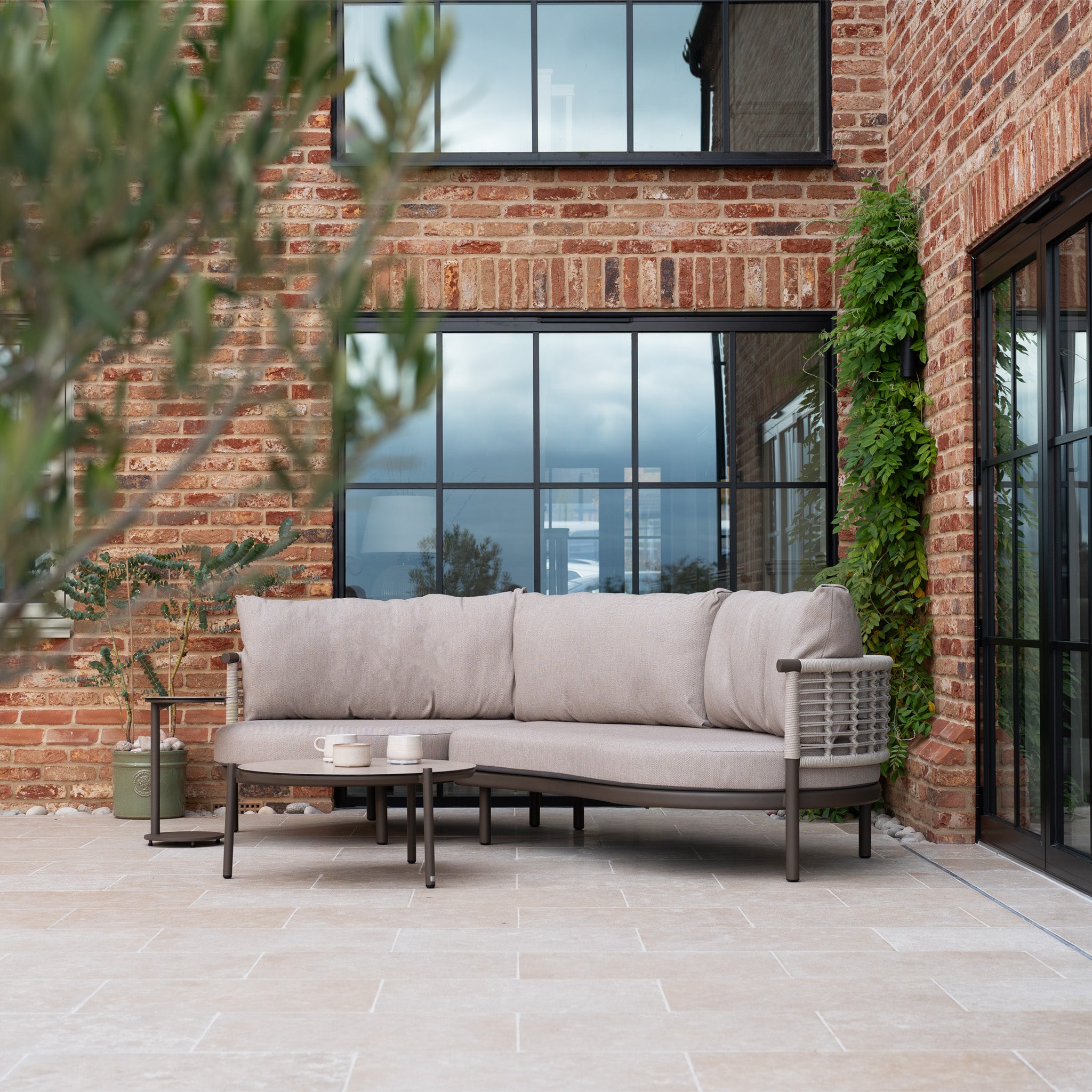 Modern outdoor patio featuring a Sutera Curved Chaise Sofa Set in Putty with all-weather cushions, a round coffee table, and brick walls with large windows.