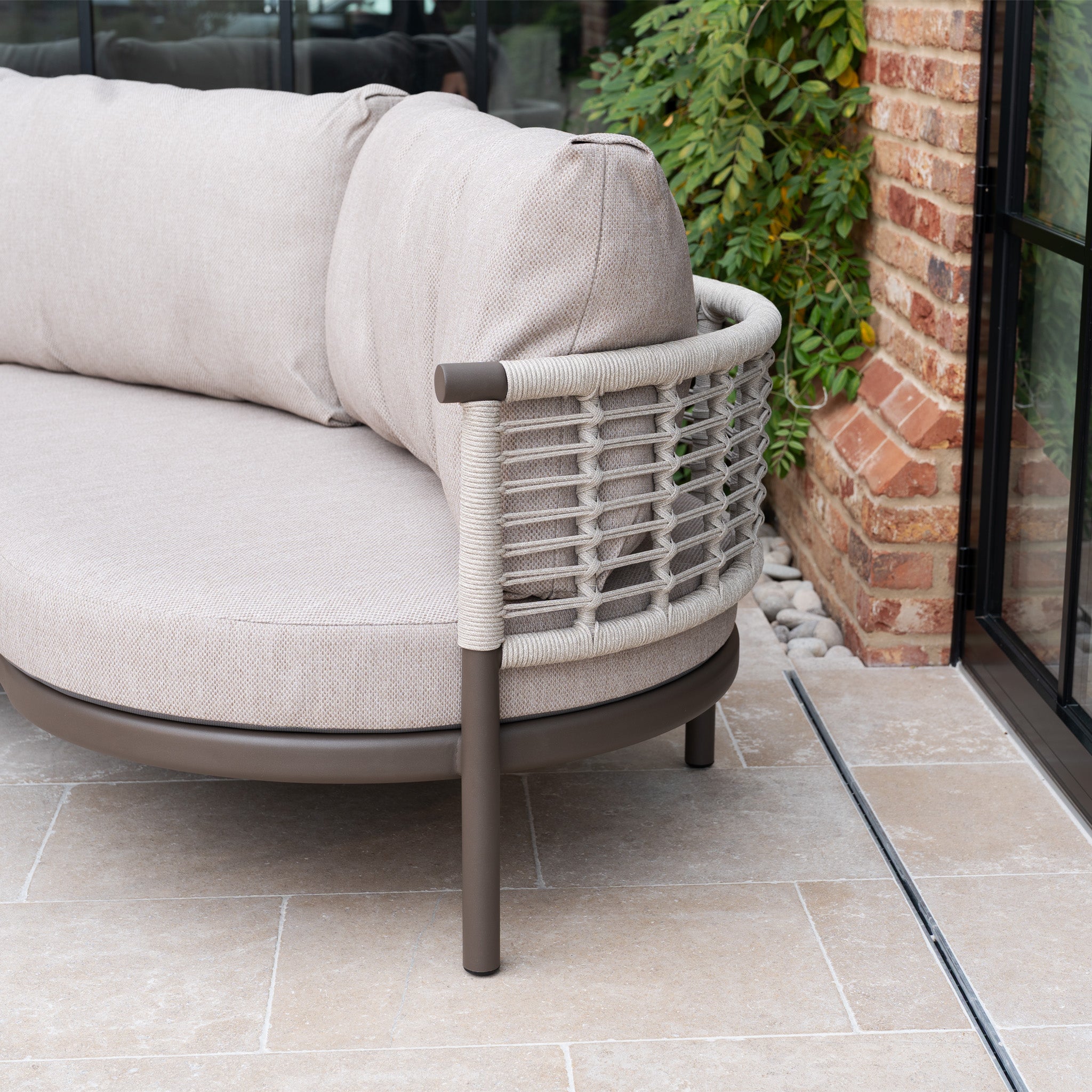 The Sutera Curved Chaise Sofa Set in Putty features woven rope details and all-weather cushions, shown on a patio beside a brick wall and vibrant green foliage.